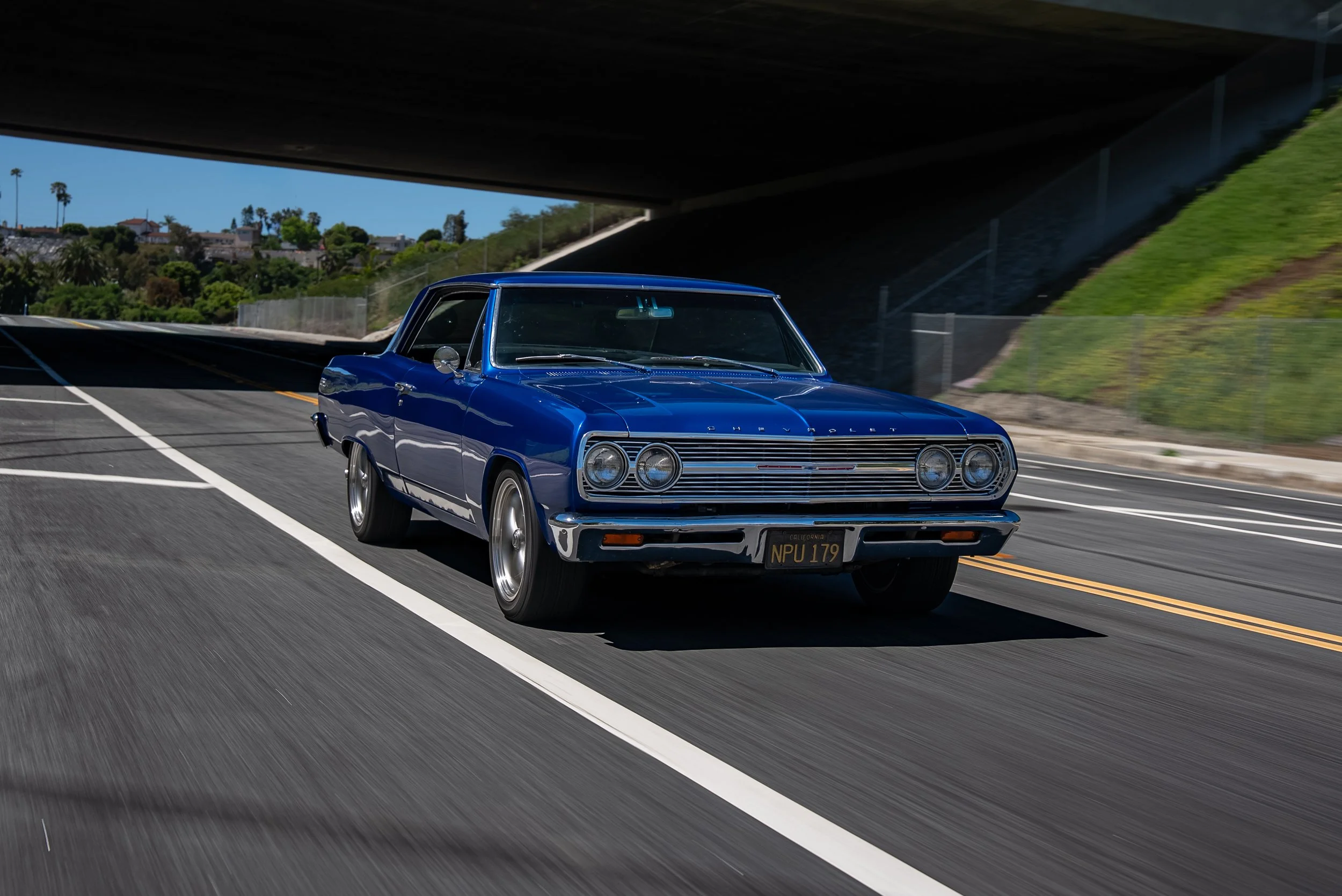 A classic blue Chevrolet car driving under a bridge on a city street.