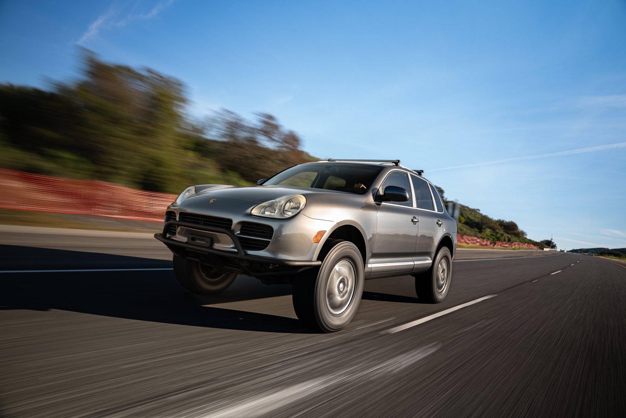 A silver Porsche SUV driving quickly on an open road with a blurred background, trees on the side, and a blue sky overhead.