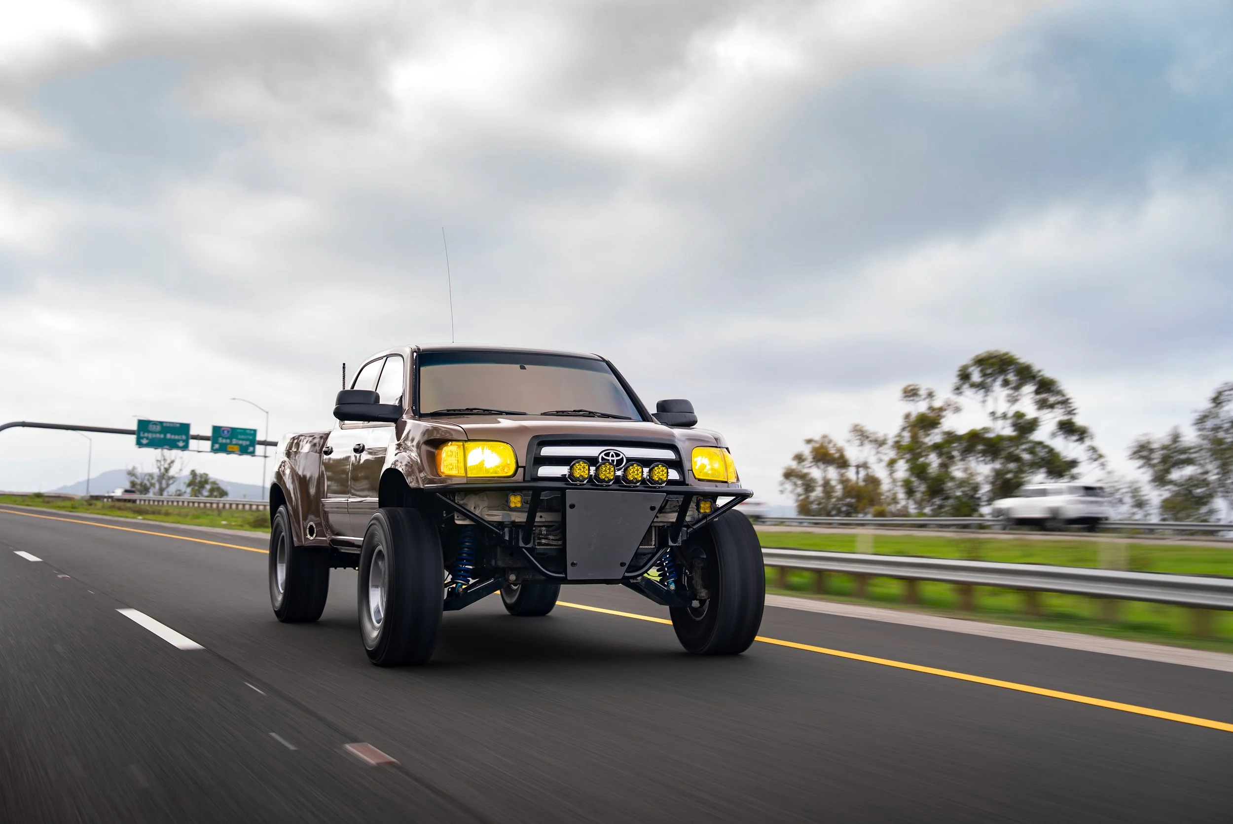 A modified Toyota pickup truck with large tires and off-road lights driving on a highway under a cloudy sky.