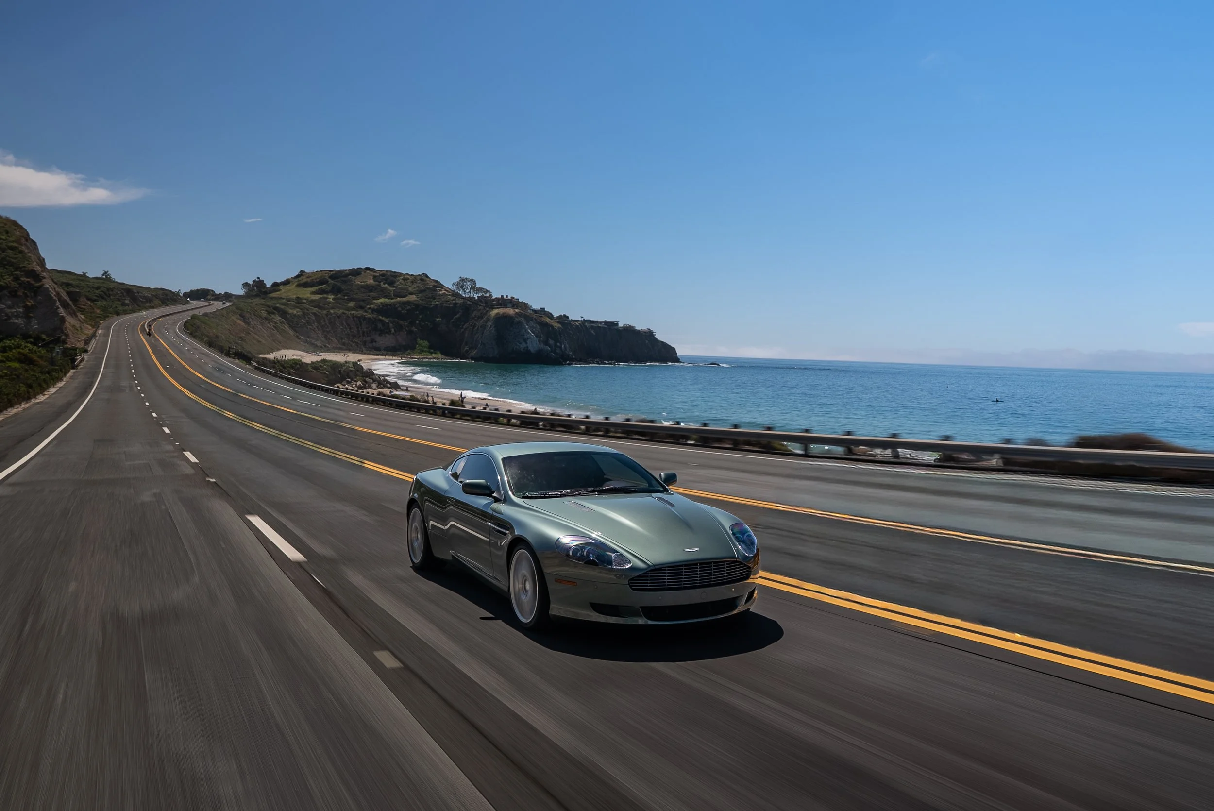 A black luxury sports car is driving on a coastal highway with the ocean and cliffs in the background under a clear blue sky.