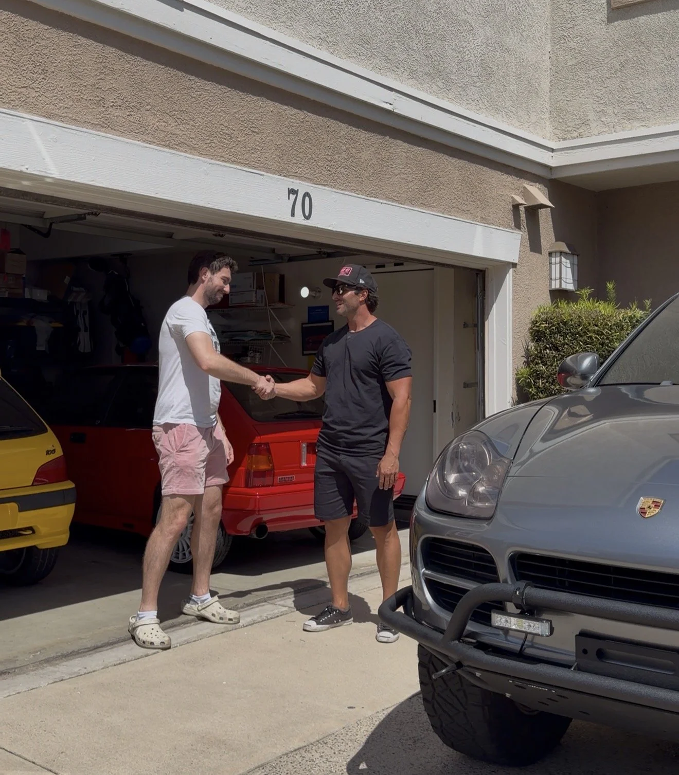 Two men shaking hands in front of a garage with multiple cars, including a yellow, red, and gray Porsche.