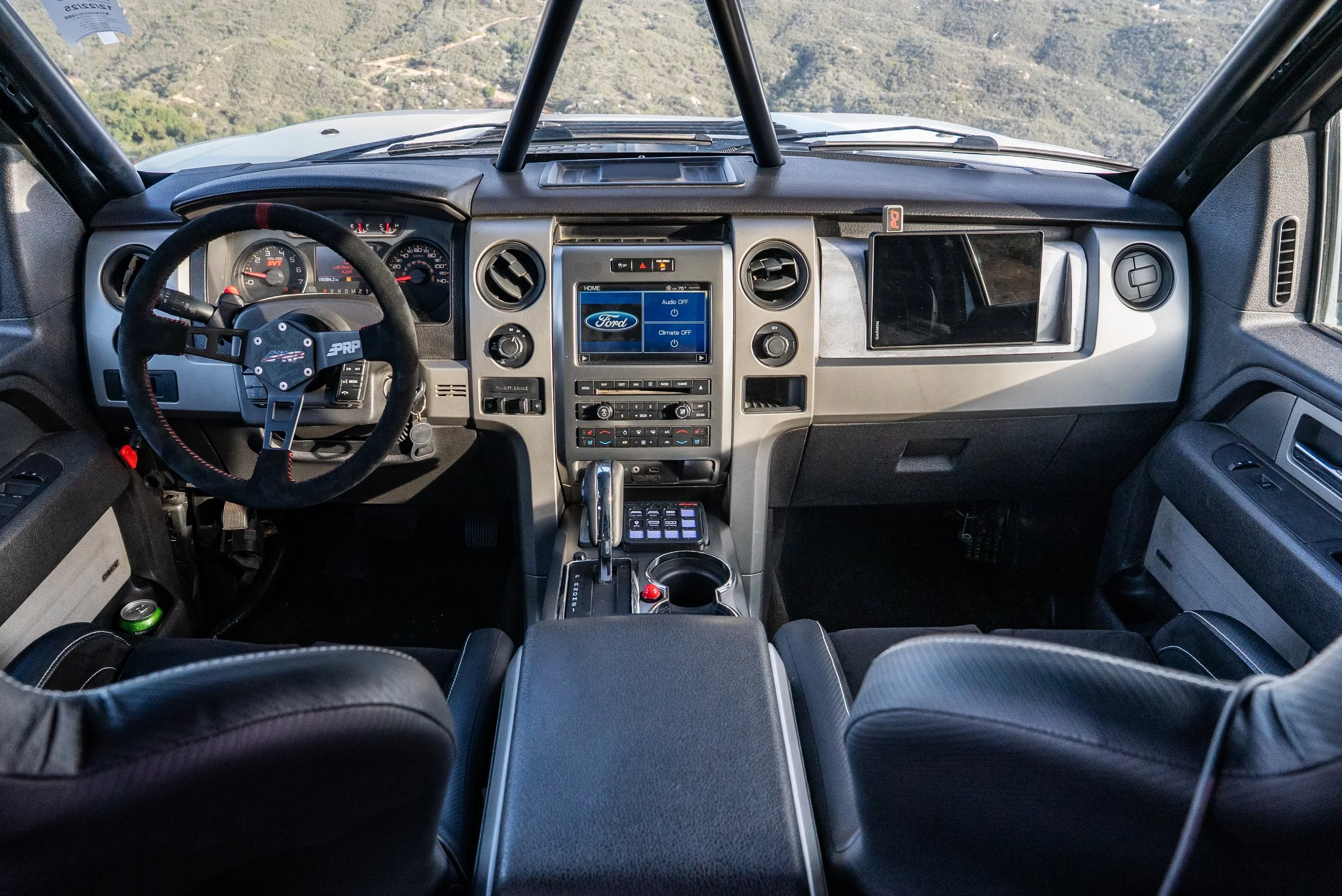 Inside view of a truck's dashboard with steering wheel, touchscreen display, gear shift, and navigation system, overlooking a mountainous landscape.