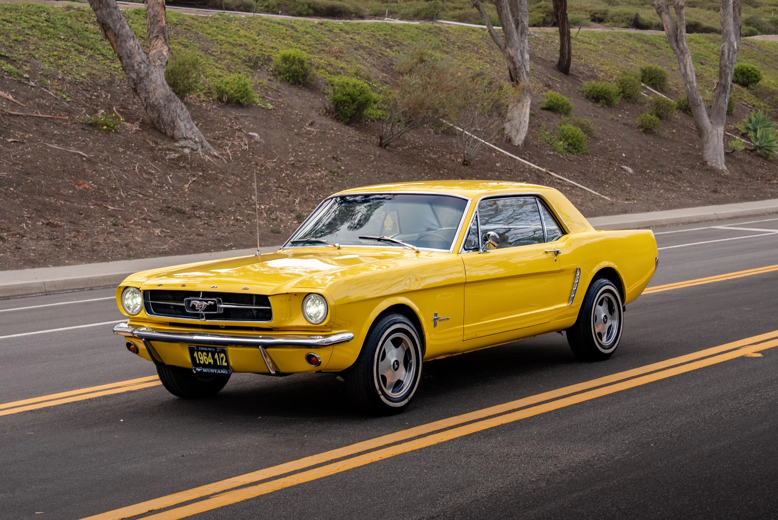 A classic yellow Ford Mustang car driving on a road with a hillside and trees in the background.