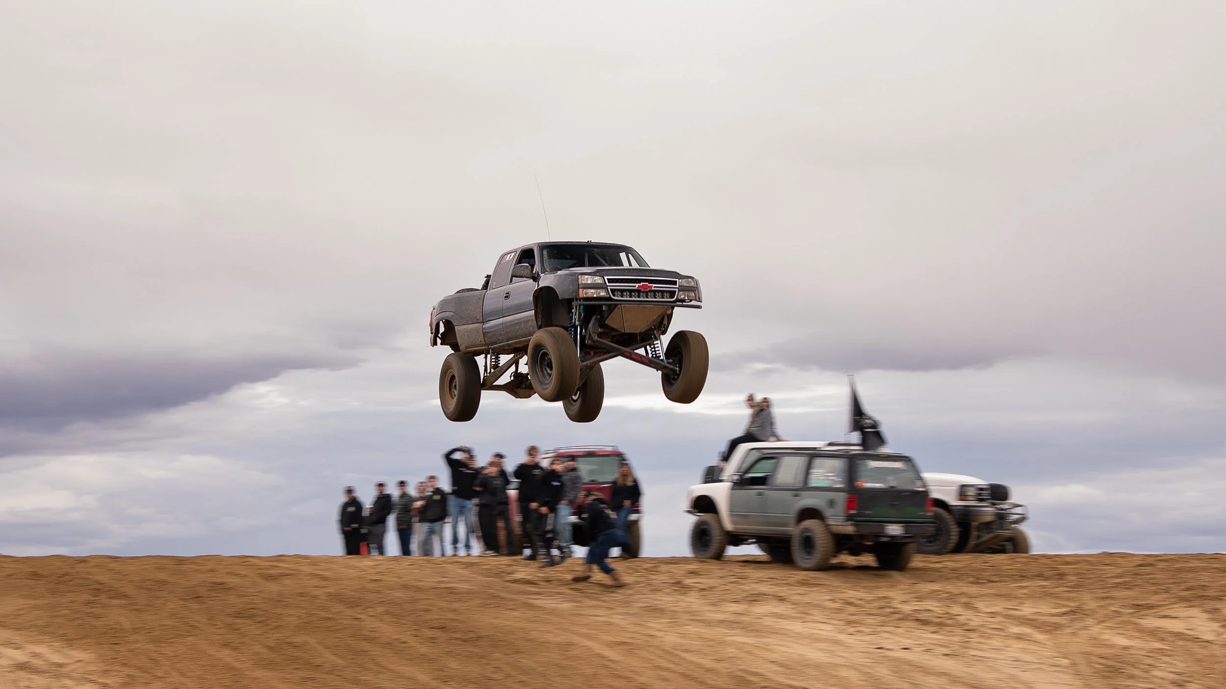 A pickup truck with large off-road tires is airborne during a jump on a dirt track, with a group of people and other vehicles in the background under an overcast sky.