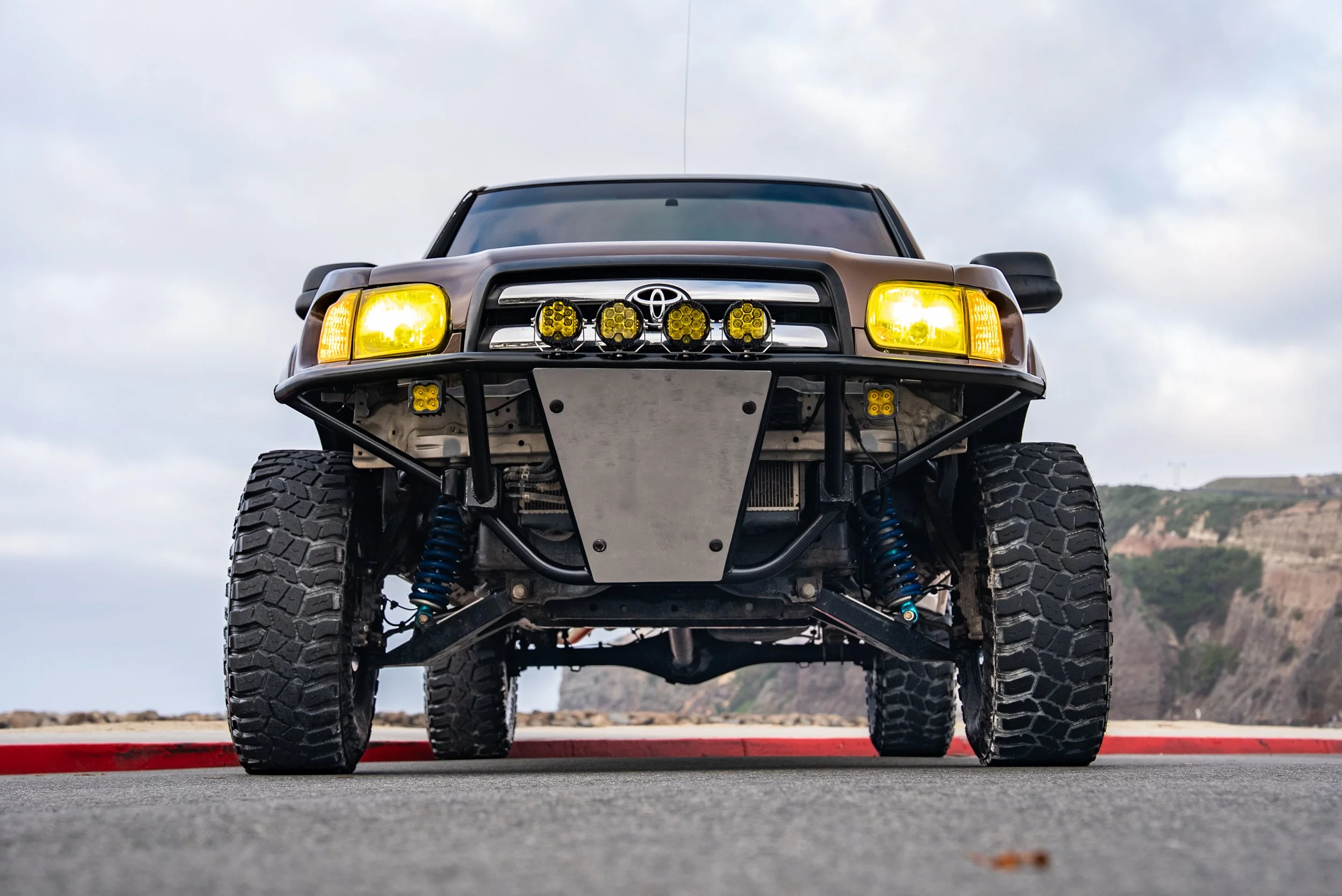 Front view of off-road vehicle with large tires, yellow headlights, and additional yellow lights on the grille, parked on a paved surface with a cloudy sky and scenic cliffs in the background.