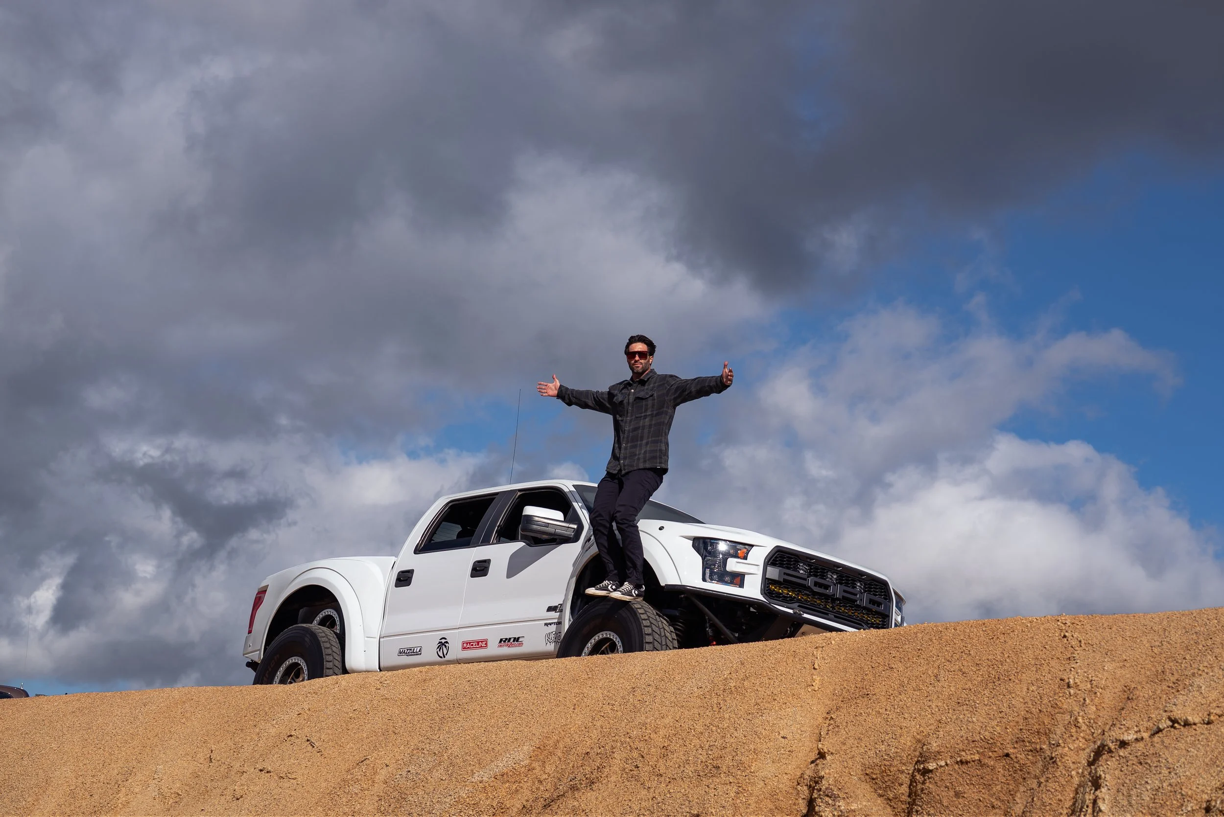 A man standing on the front of a white truck with open arms, on a dirt mound, against a cloudy sky.