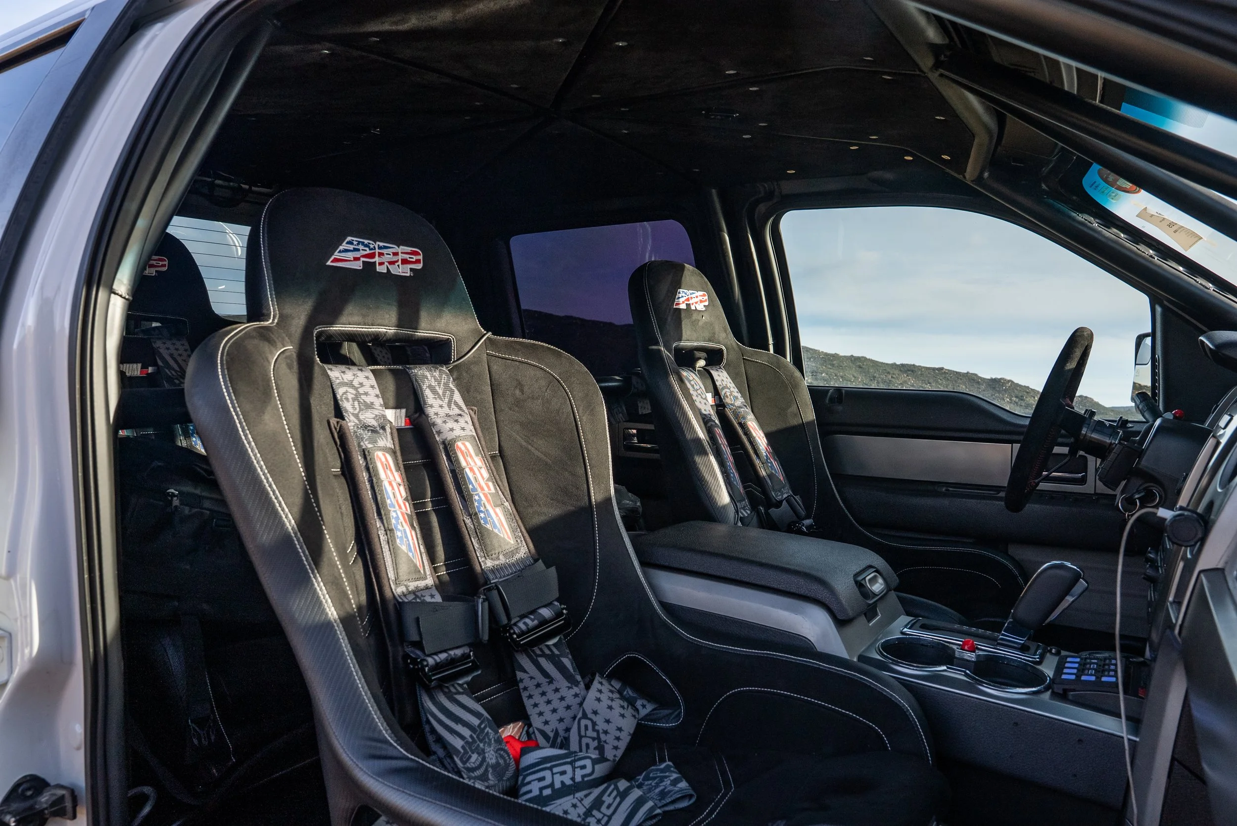 Interior of a race car with two racing seats, racing harnesses, and a dashboard.