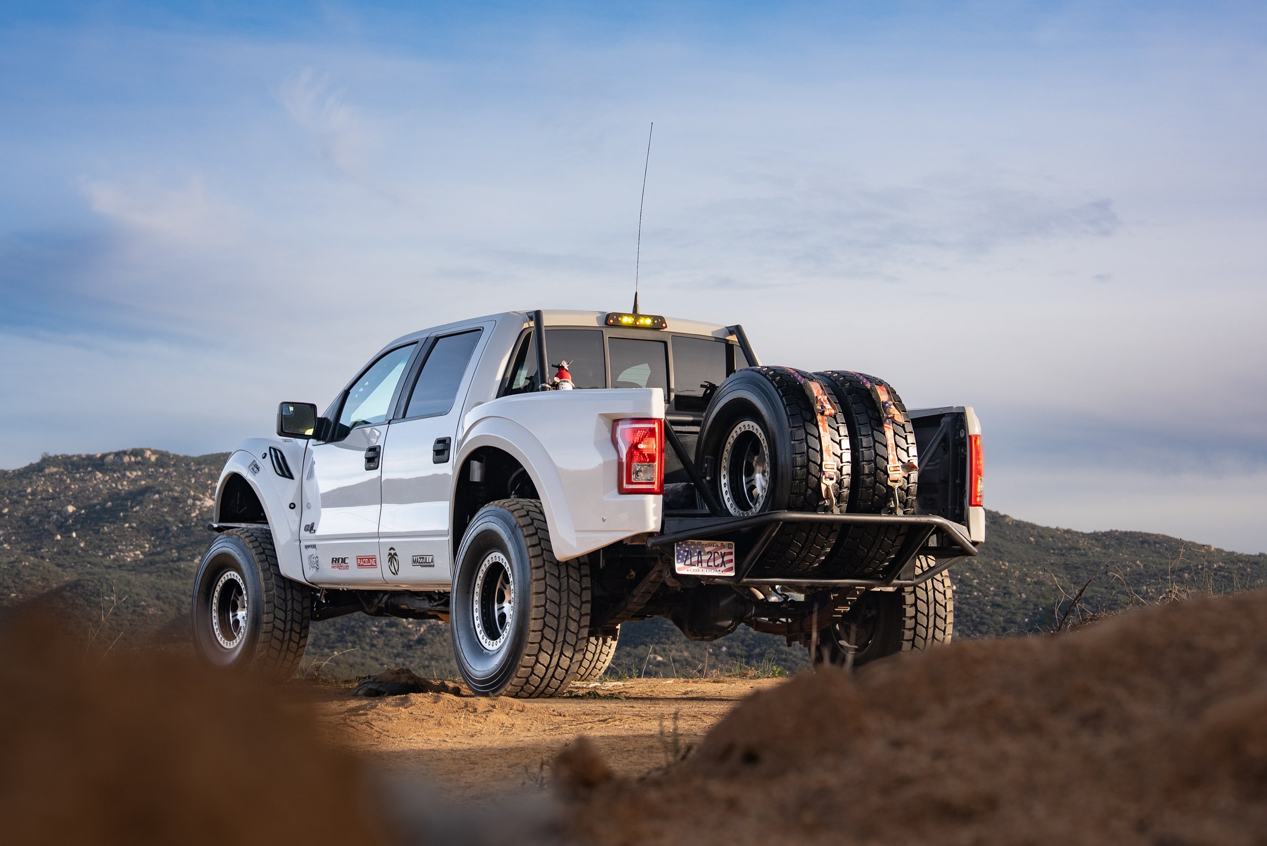 A white off-road pickup truck with spare tires mounted on the back, parked on a dirt trail with hills in the background.