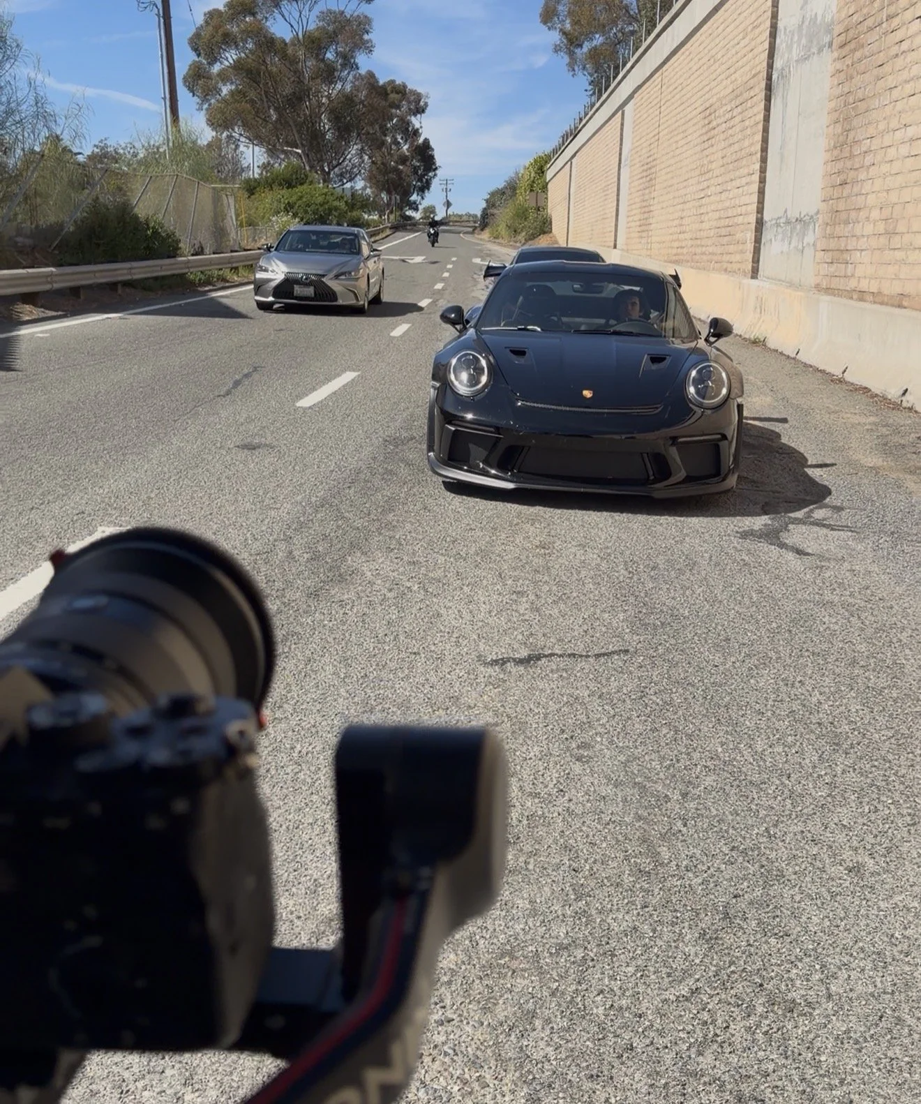 Multiple cars on a slope street, including a black sports car in the foreground and a grey sedan in the background, with trees and a brick wall alongside the road.