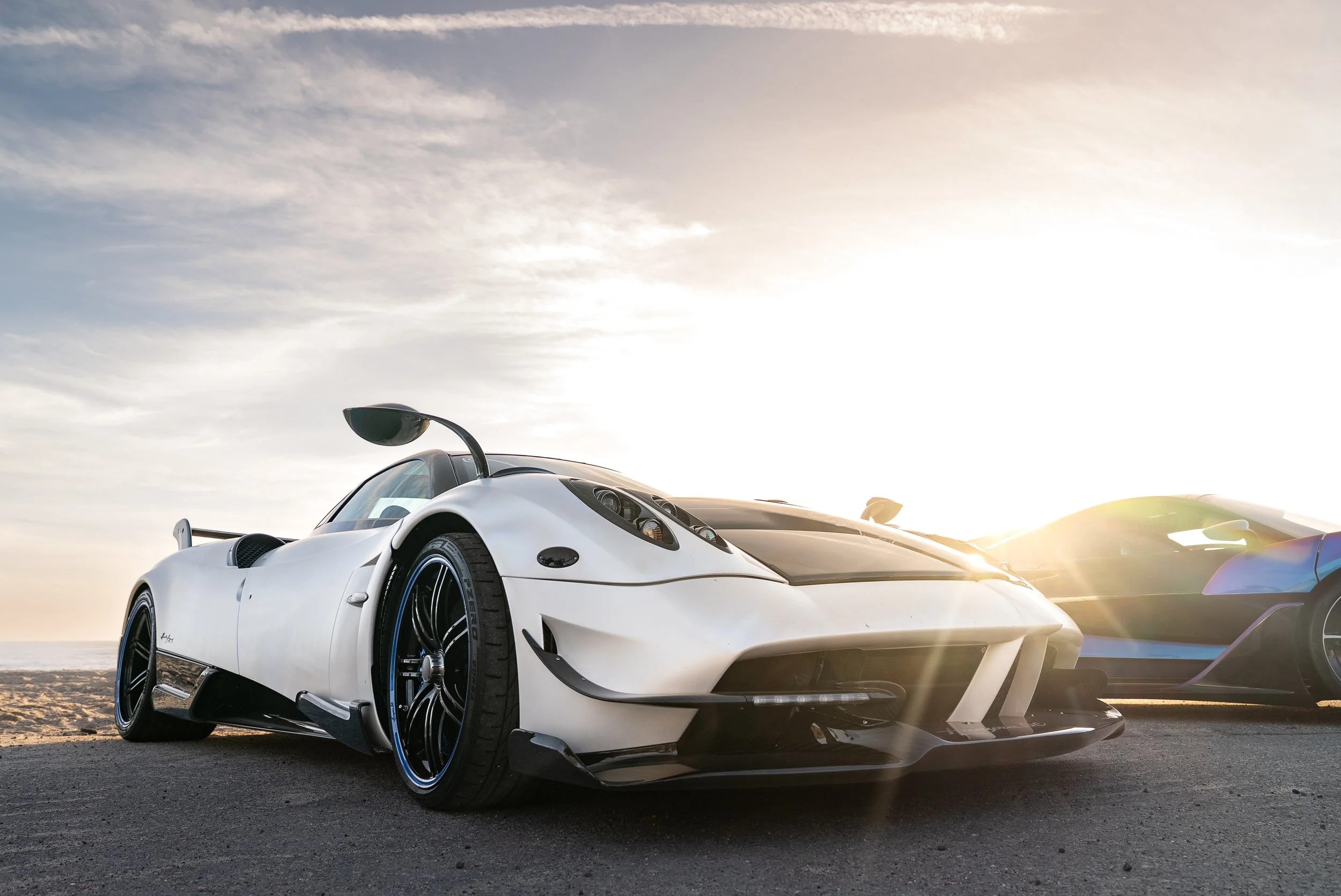 A white sports car parked on the road during sunset with another sports car and the ocean in the background.