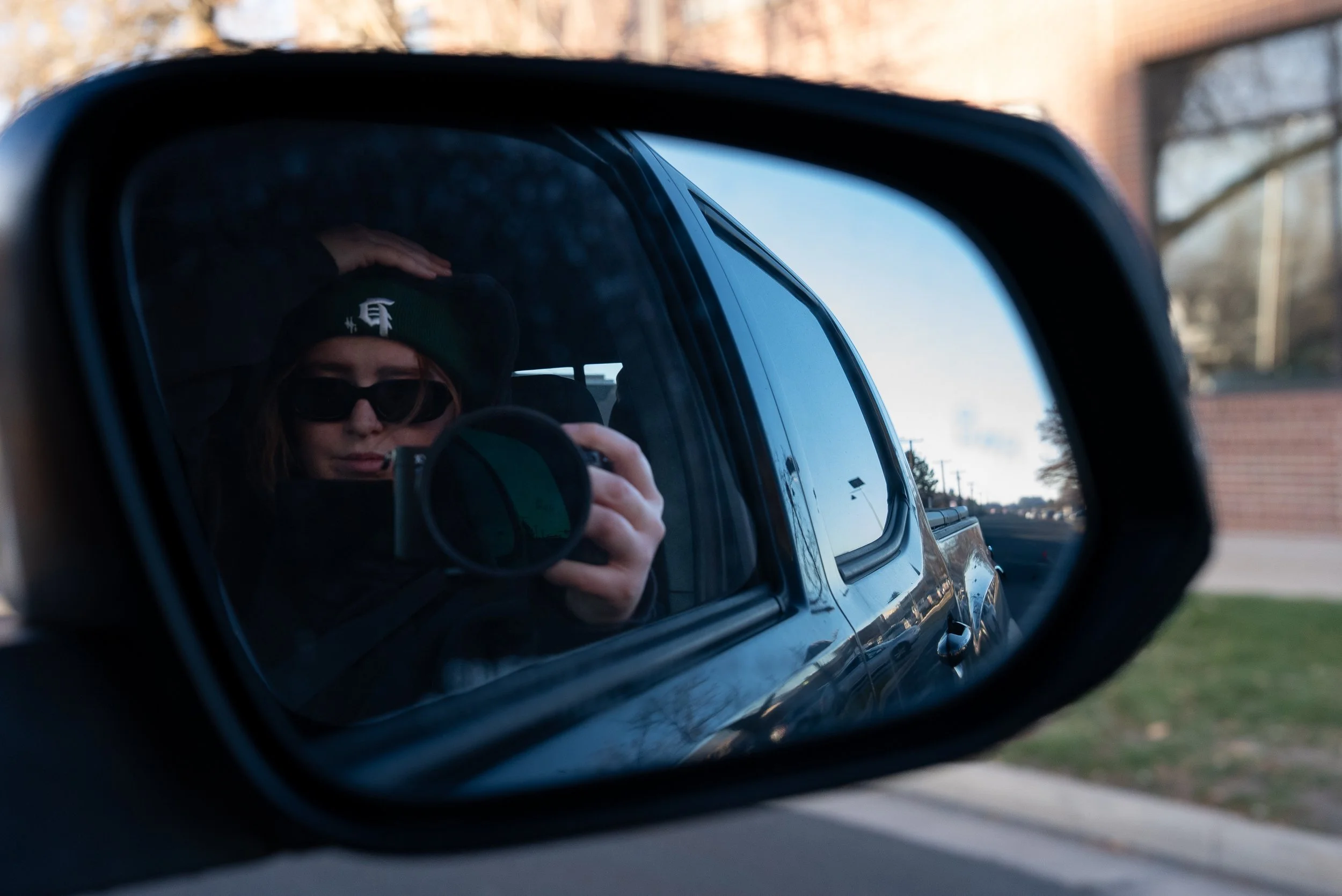 Person taking a photo of themselves in a car side mirror, wearing sunglasses and a black beanie with a white logo.