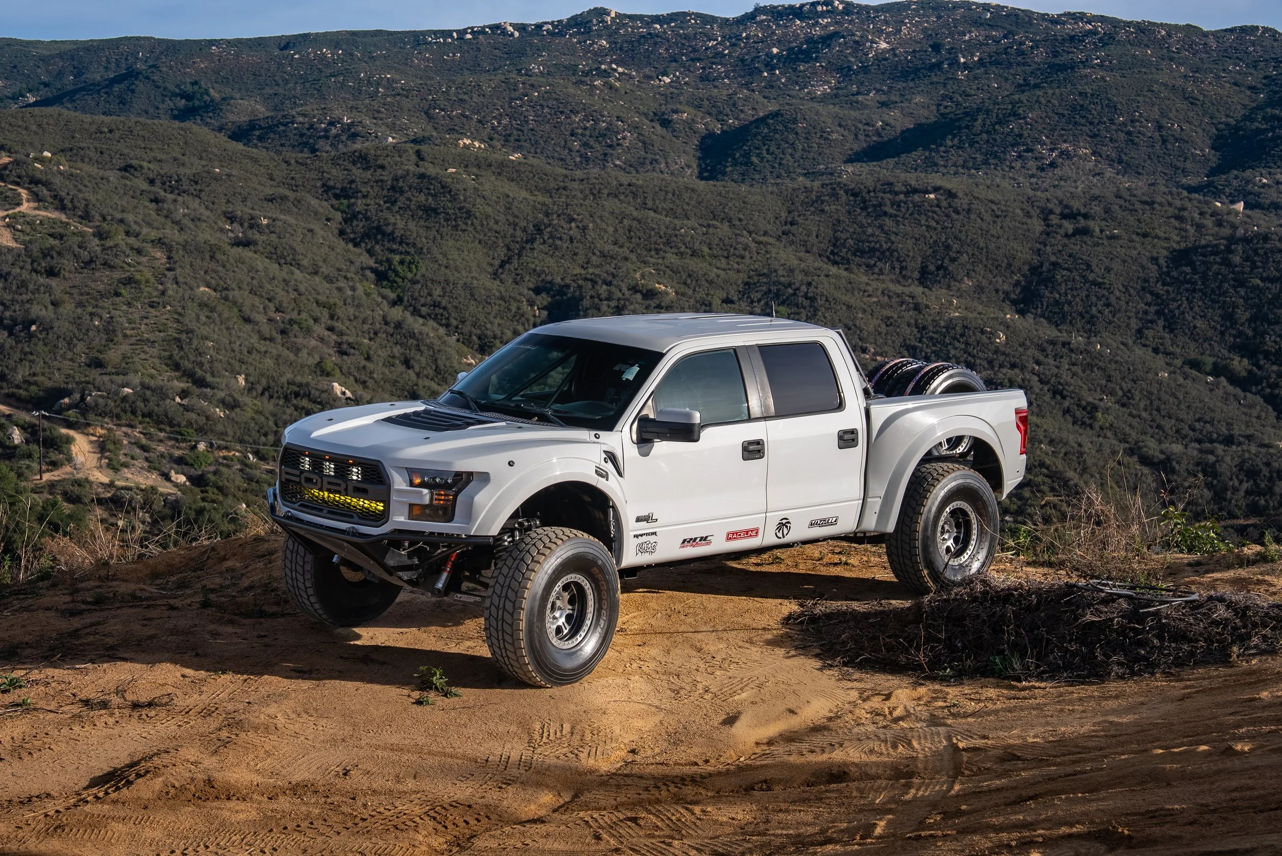 A white off-road pickup truck with modifications parked on a dirt trail in a hilly, green landscape.