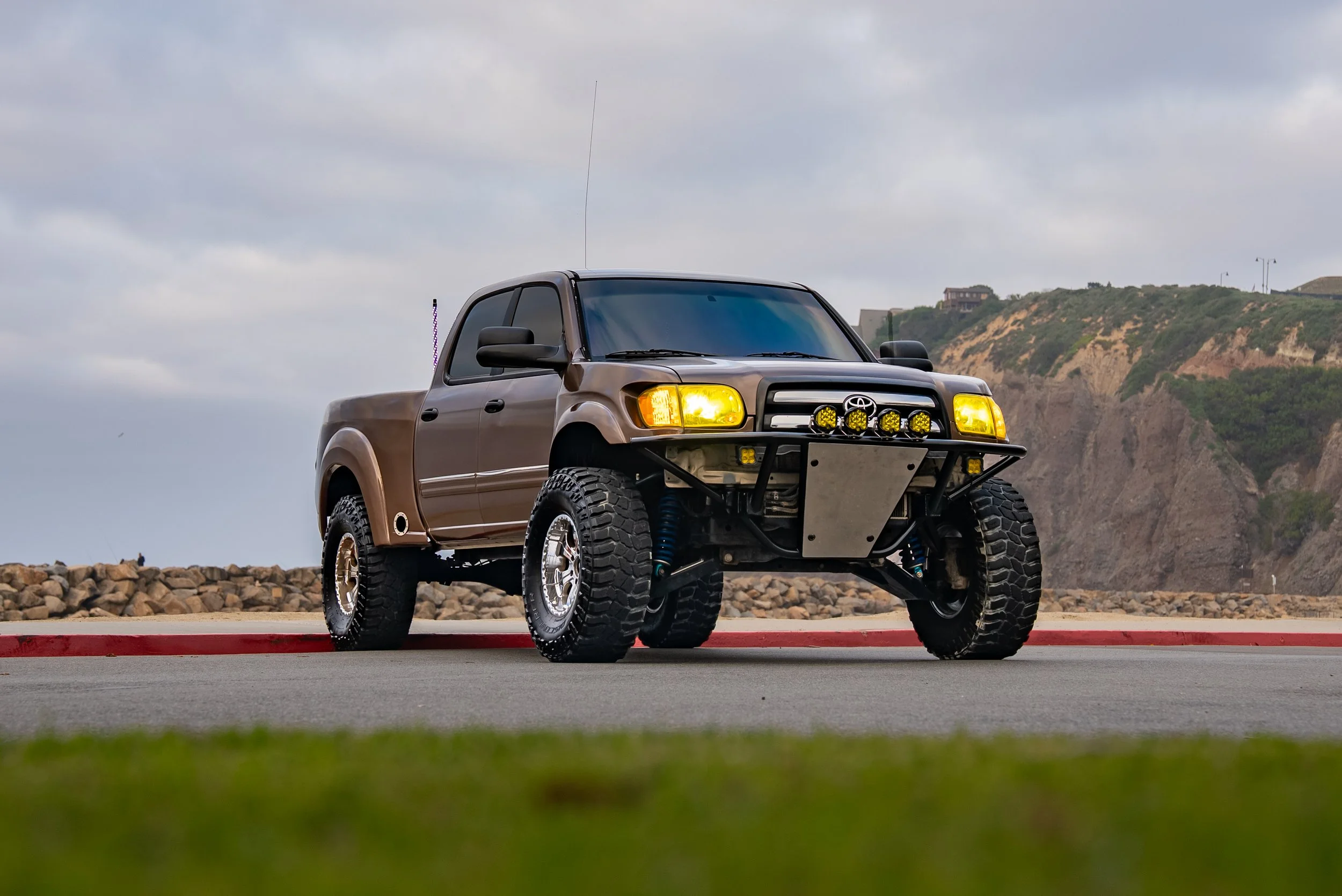 A modified brown Toyota Tacoma pickup truck with off-road tires, custom front bumper, and yellow headlights parked near a coastline with rocky cliffs in the background.