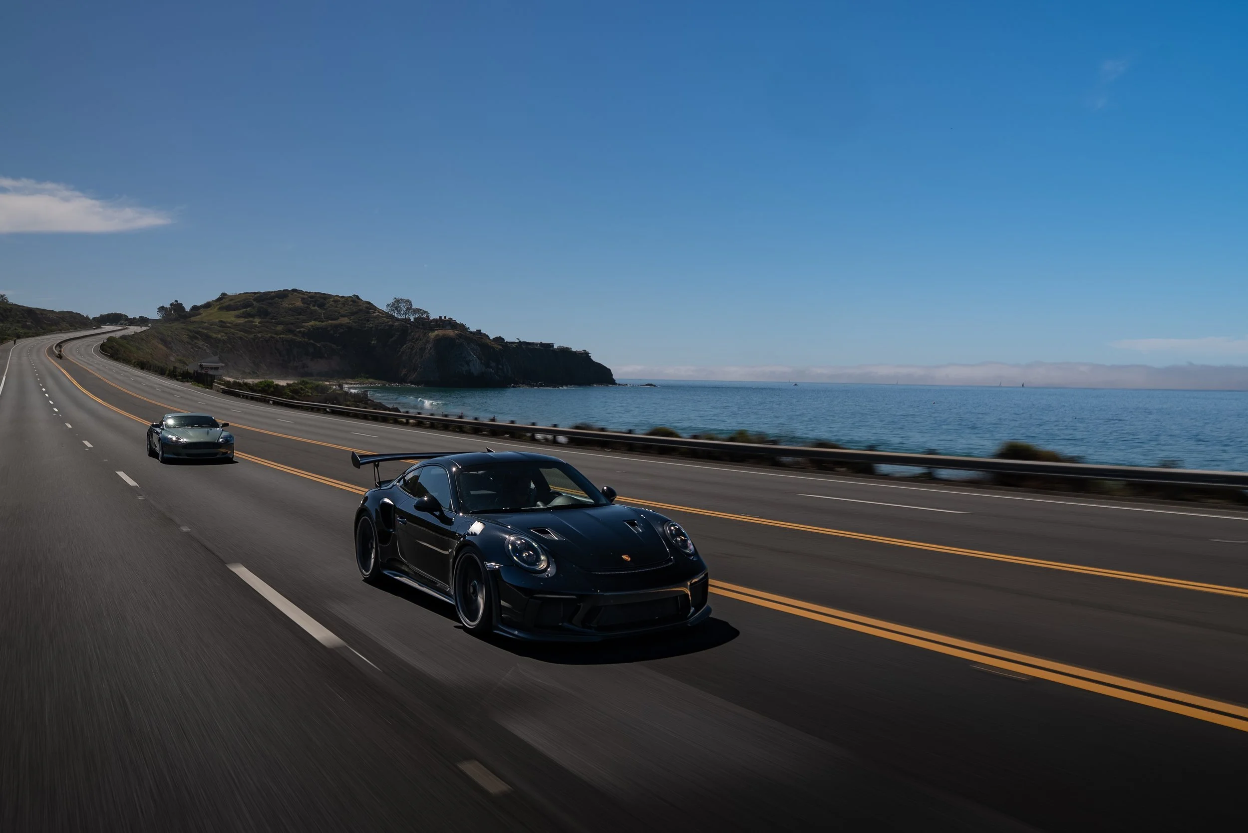A black Porsche race car driving on a coastal highway with two other cars, near a scenic ocean view with blue sky and hills in the background.