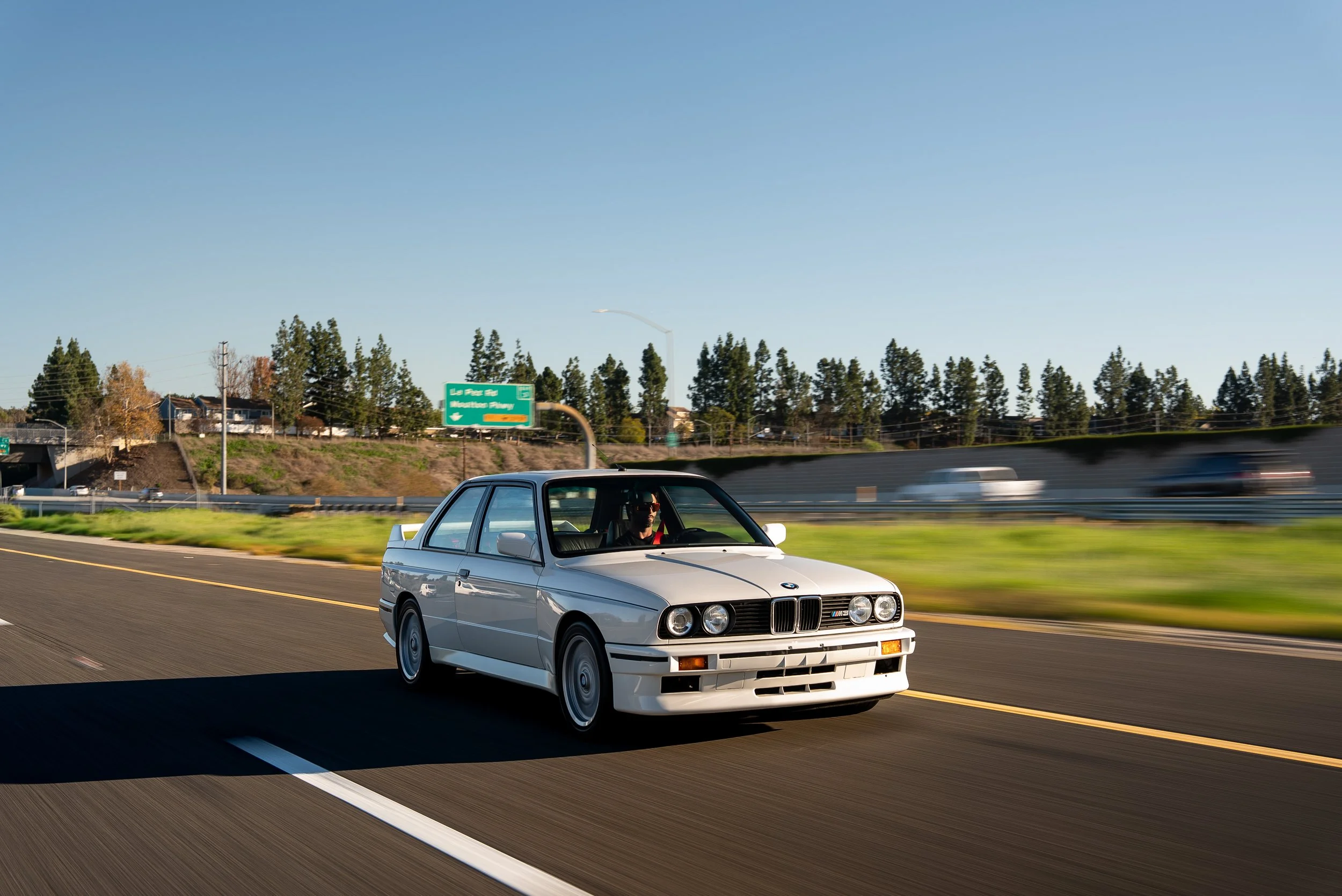 A white vintage BMW car driving on a highway with a person inside, green trees and highway signs in the background, under a clear blue sky.