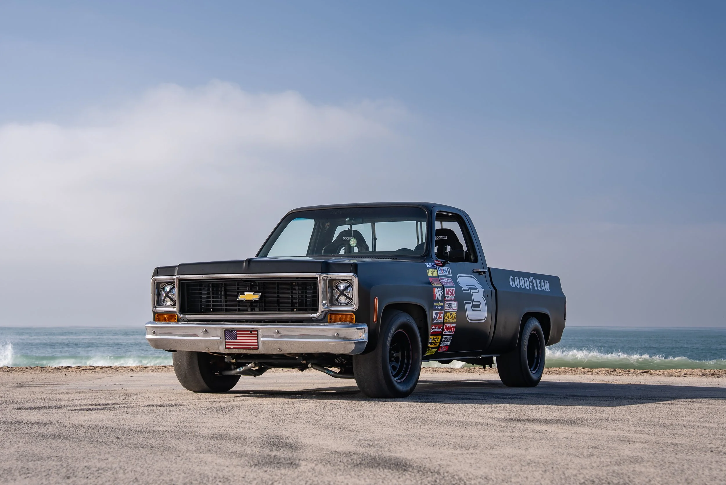 A vintage black Chevrolet pickup truck with racing decals and the number 3 is parked on a sandy beach with the ocean and cloudy sky in the background.