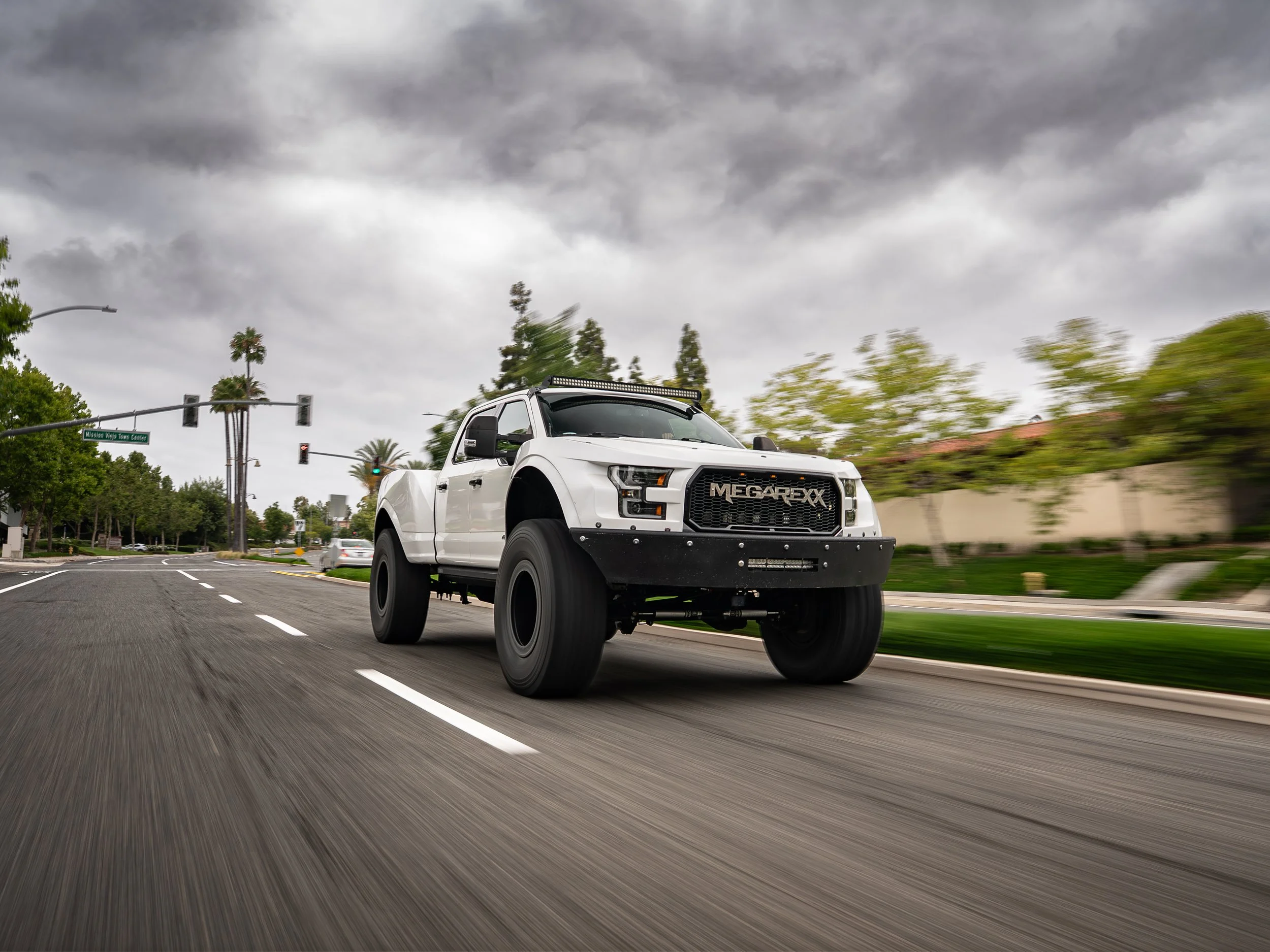 A white off-road pickup truck with black wheels and MEGARACX branding on the front grille driving on a city street under cloudy skies.