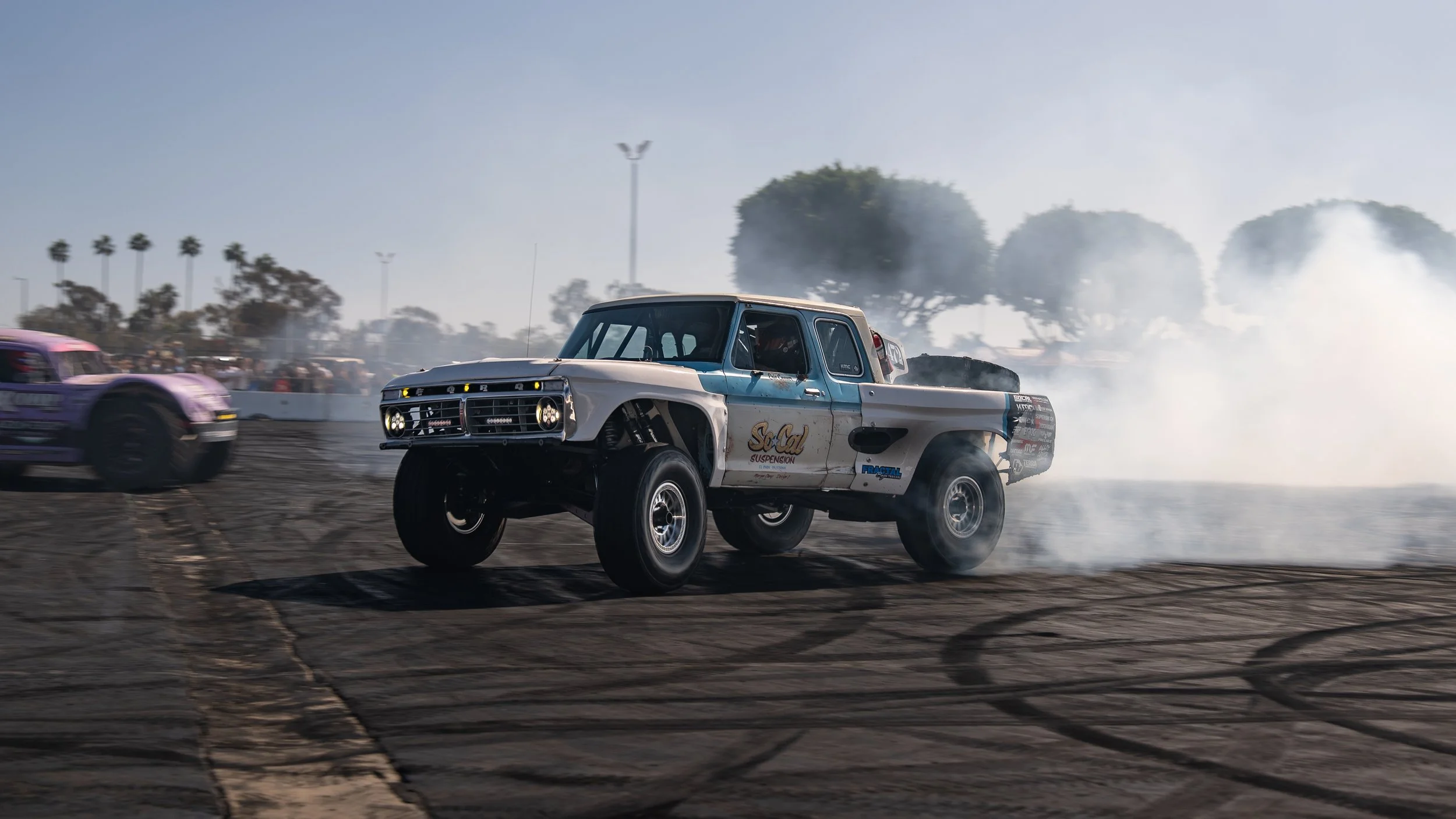 A vintage off-road racing truck drifting while smoke billows behind it during a race, with a purple race car nearby and trees in the background.
