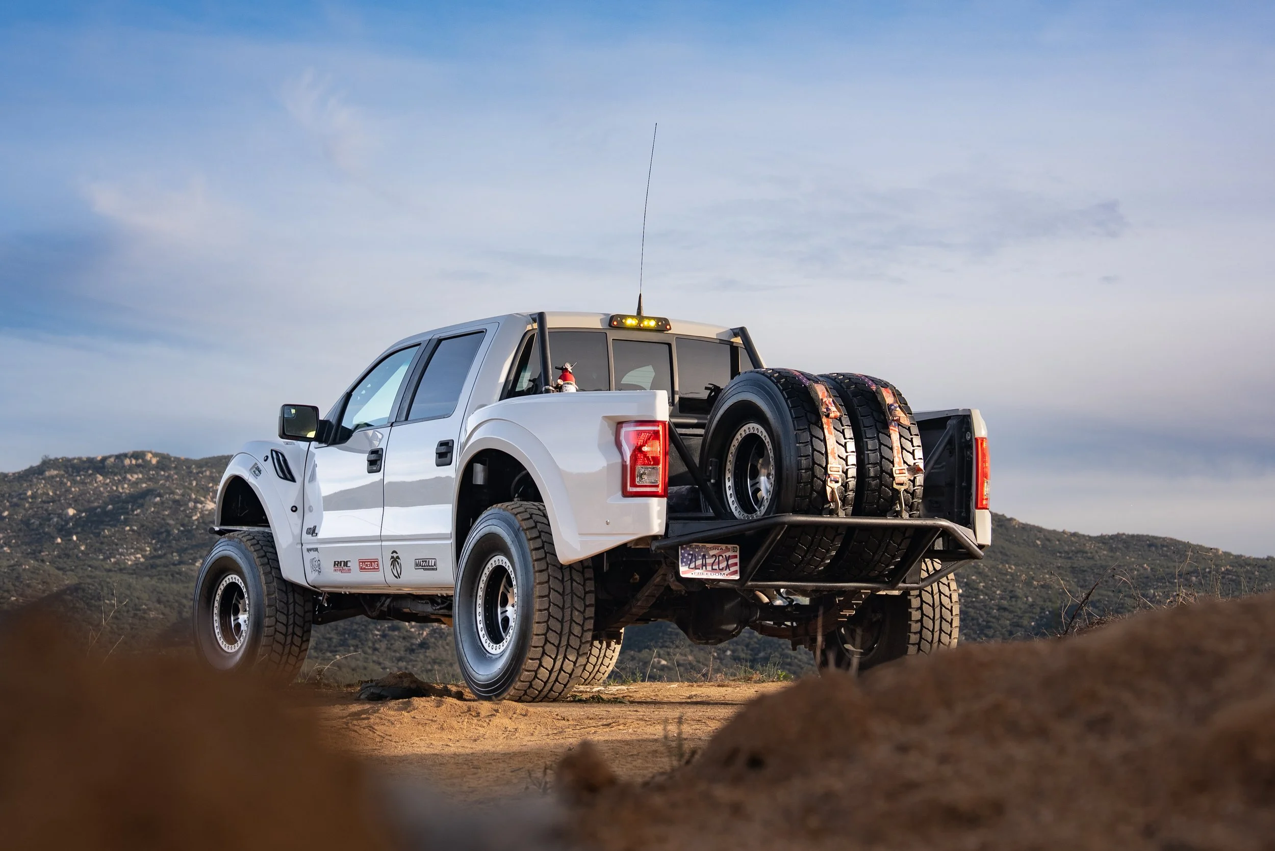 White off-road pickup truck with spare tires in the truck bed, parked on dirt with hills in the background and a partly cloudy sky.