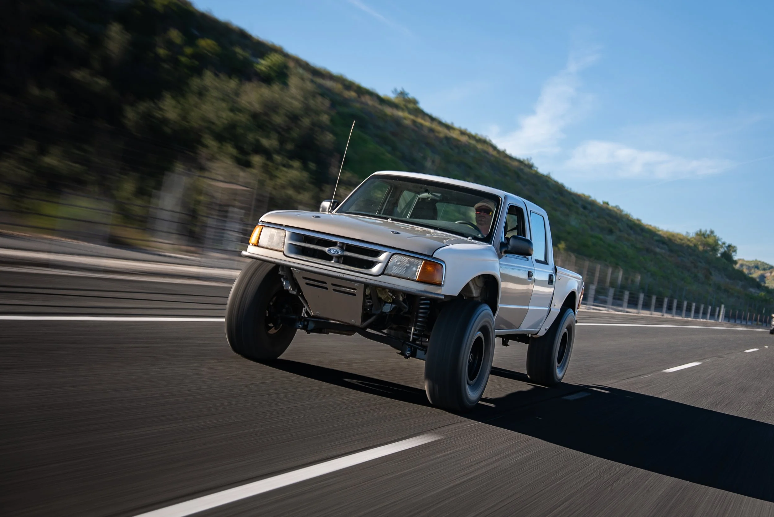 A silver Ford pickup truck with enlarged tires driving at high speed on a highway surrounded by green hills, the road appears blurred to show motion.