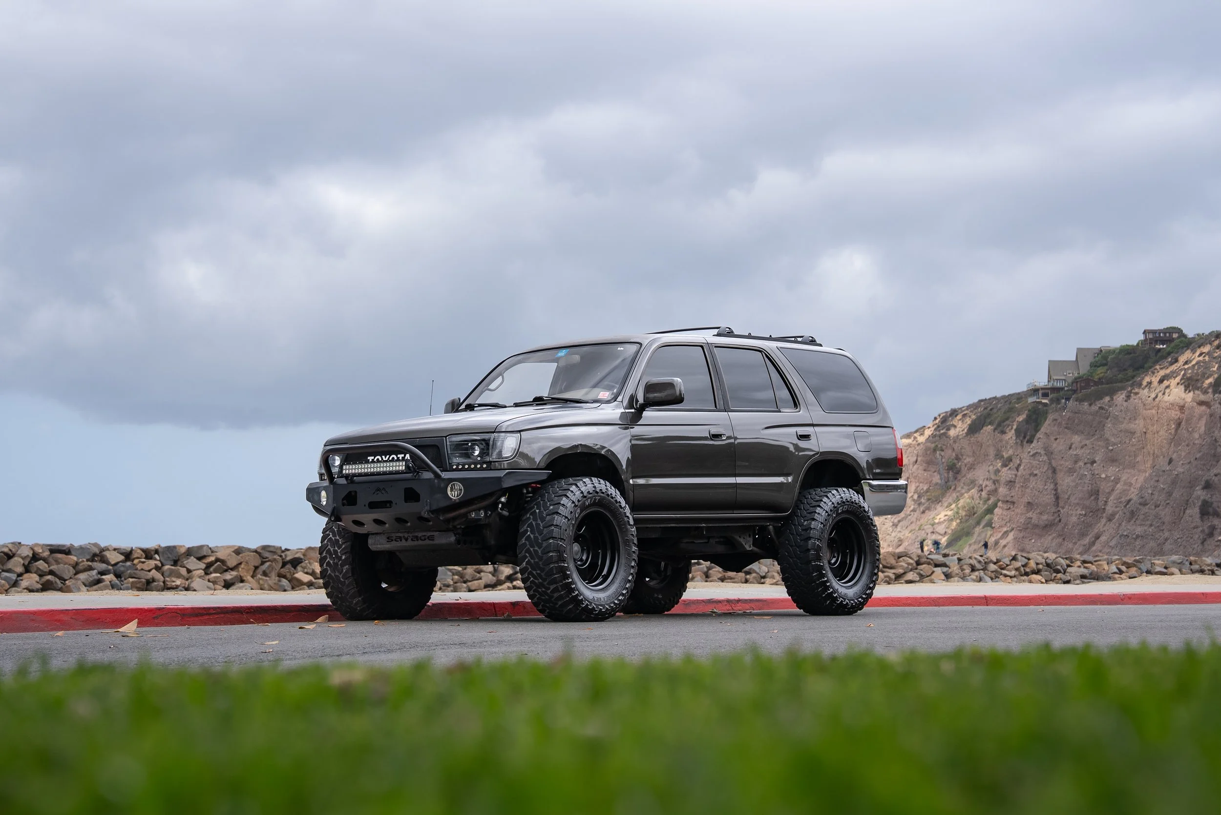 A black SUV with off-road tires and modifications parked on a coastal road with rocky shoreline and cliffs in the background.