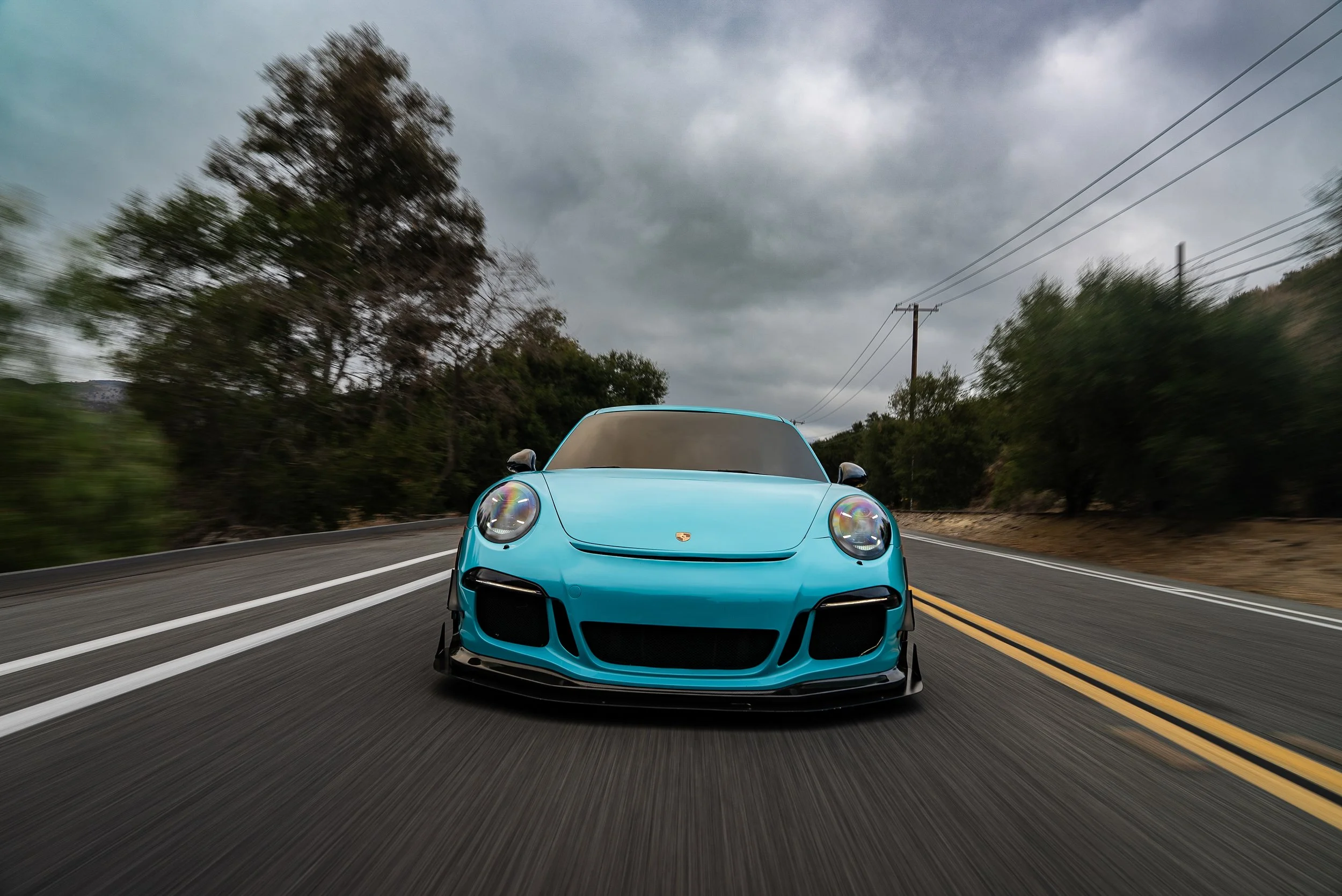 A blue sports car driving on an open road with cloudy skies and trees on the sides.
