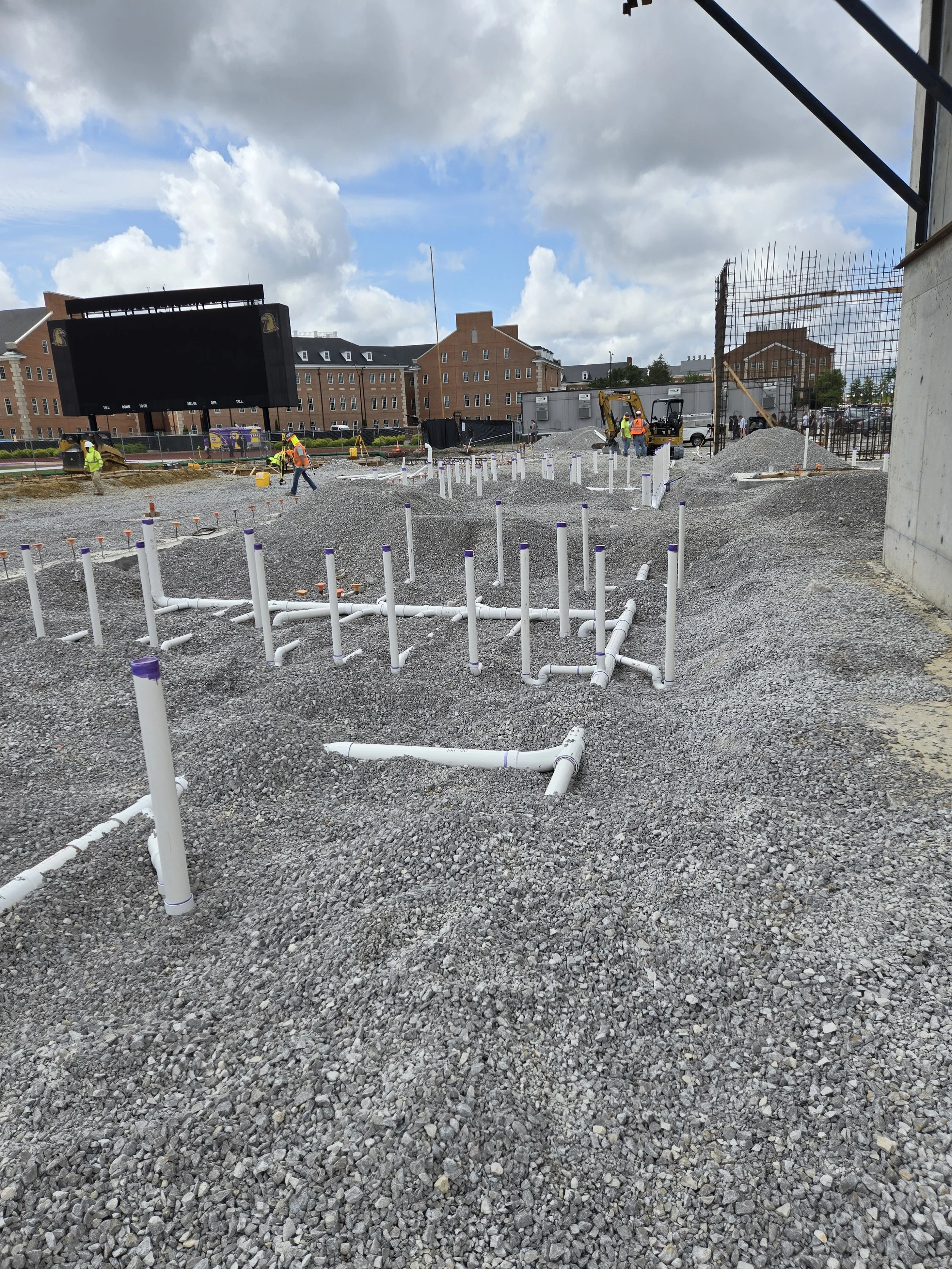 Construction site with gravel ground, white PVC pipes, and workers in safety vests and helmets; background shows significant building structure, large screen, and modern brick buildings.