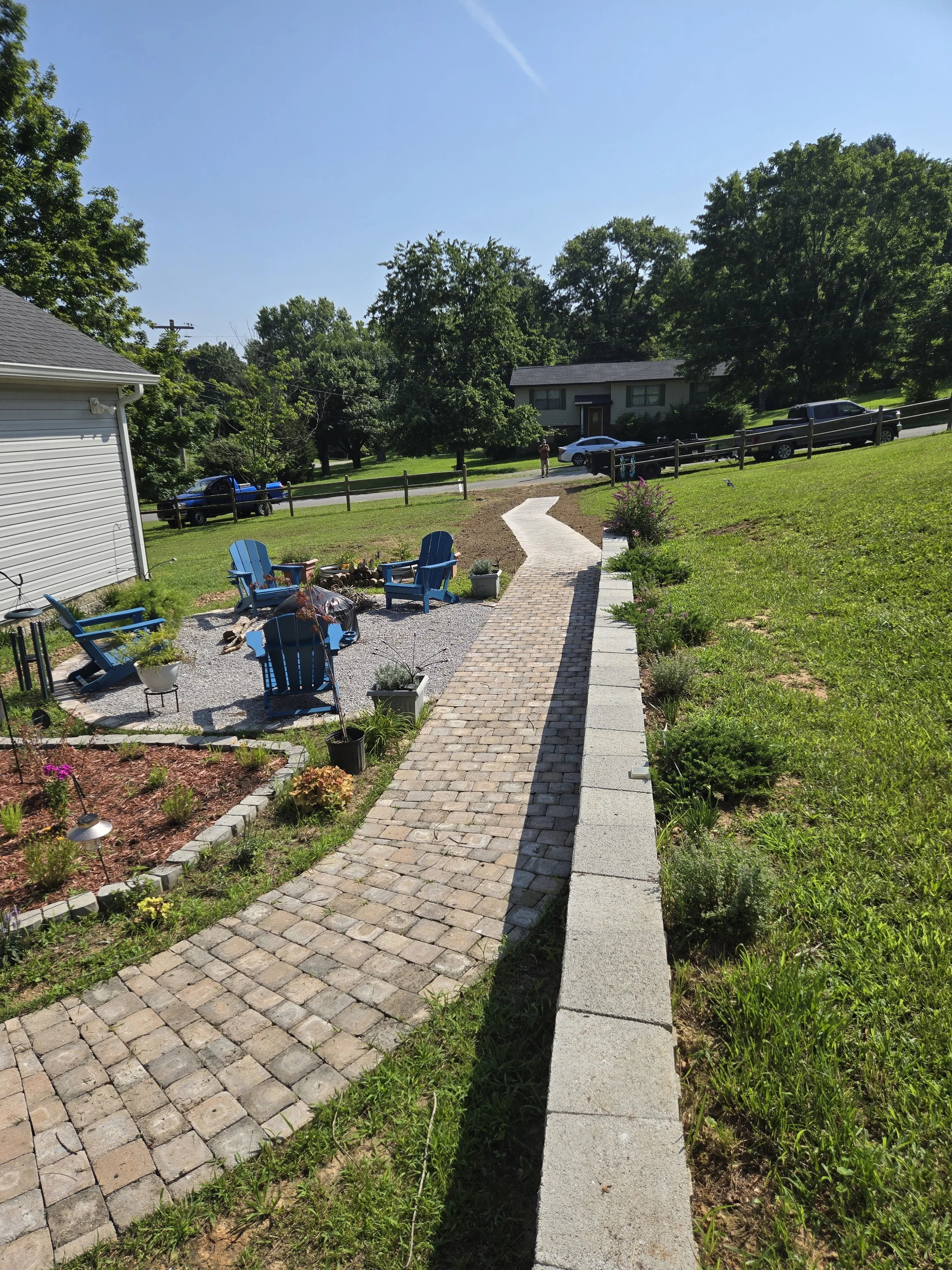 A backyard garden with a brick pathway leading to a street, surrounded by flowering plants and green grass, with trees and houses in the background on a sunny day.