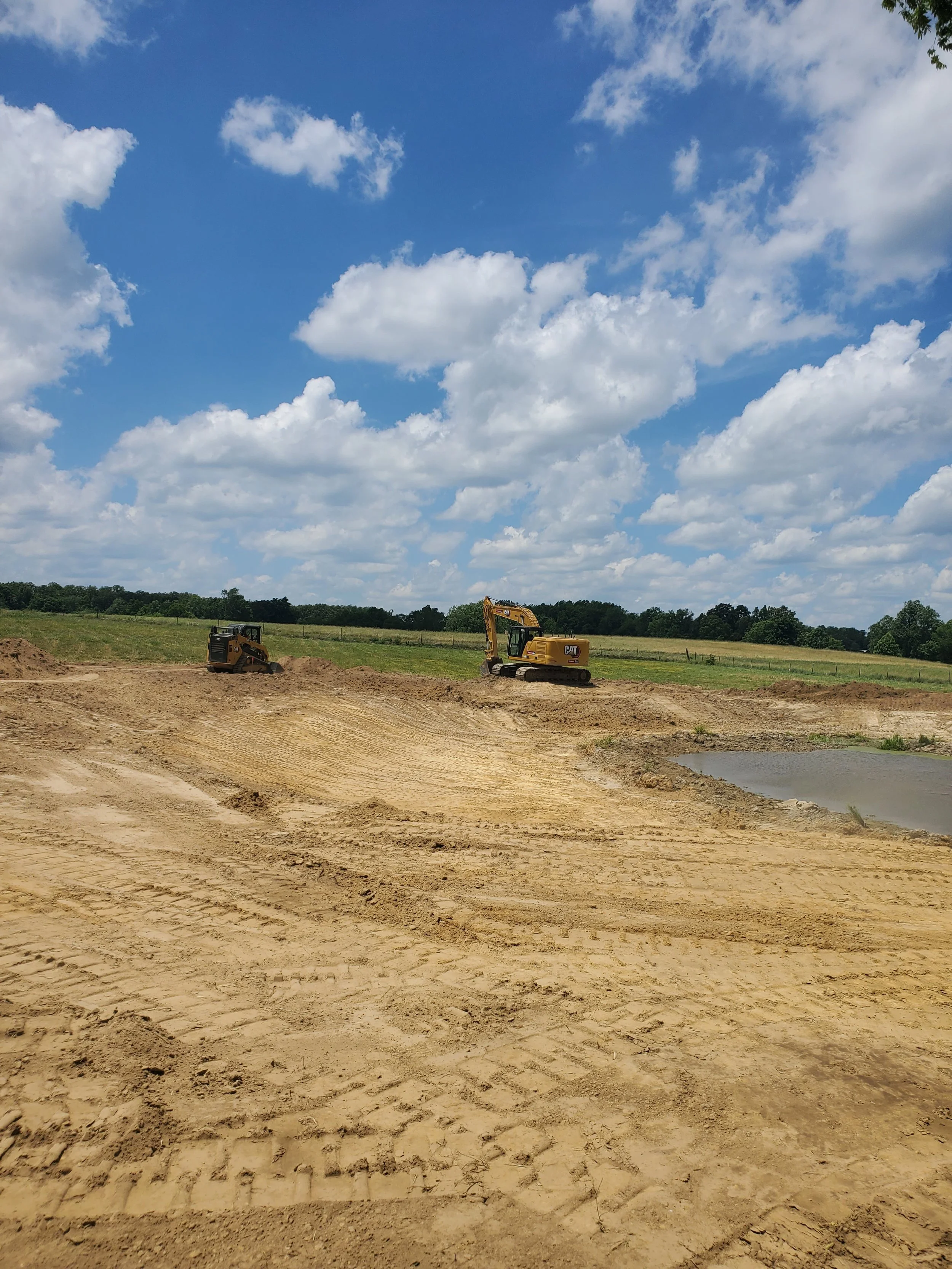 Construction site with heavy machinery, including a bulldozer and an excavator, on a dirt field under a partly cloudy sky.
