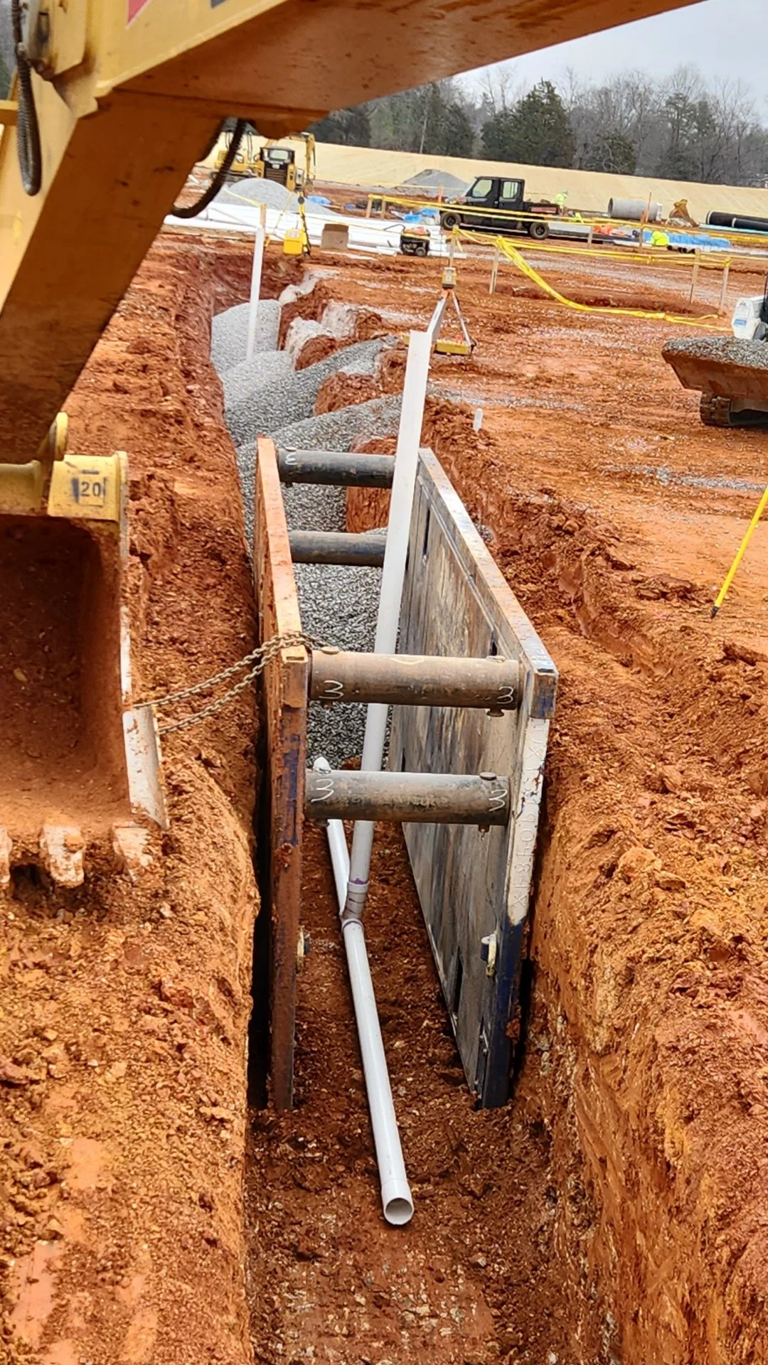 Construction site with trenches, gravel, pipes, and equipment, showing ongoing underground infrastructure work in a muddy area.