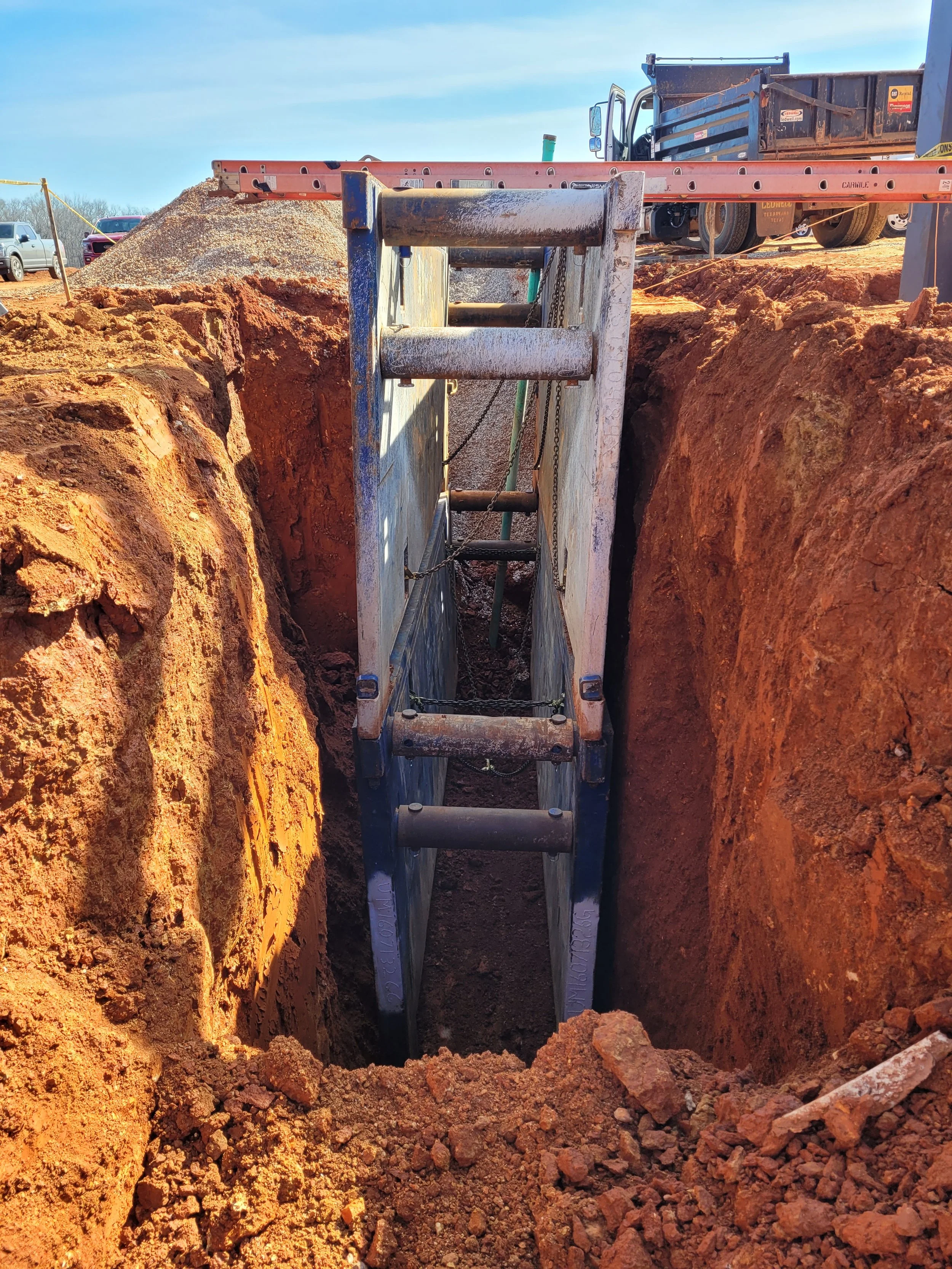 A construction site with a large, deep excavation. Inside the trench, a metal trench box supports the walls. A ladder is leaning against the trench box for access. In the background, there are construction vehicles, including a dump truck and cars, under a clear blue sky.