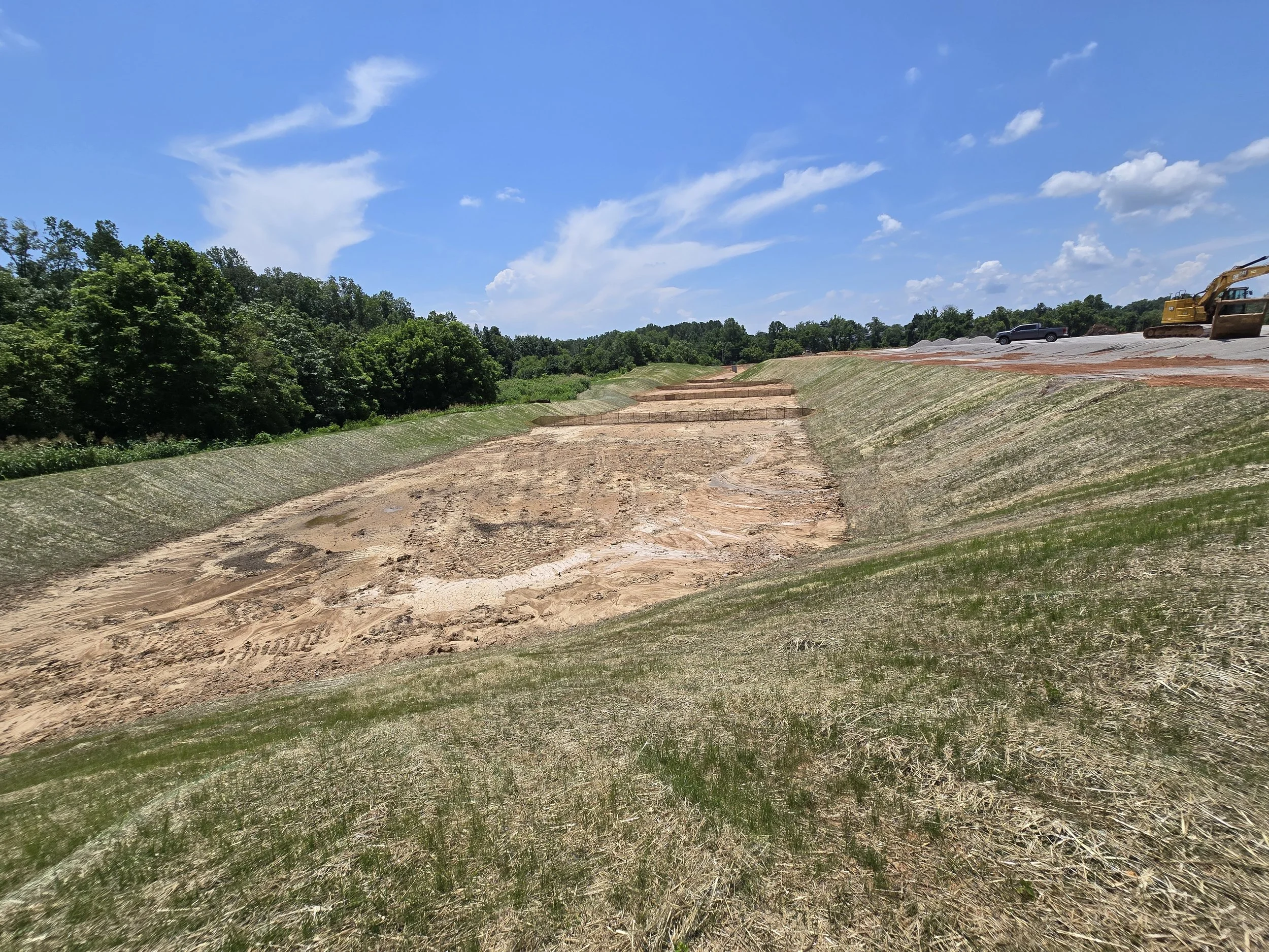 Construction site with excavator, truck, and gravel piles under a partly cloudy blue sky.