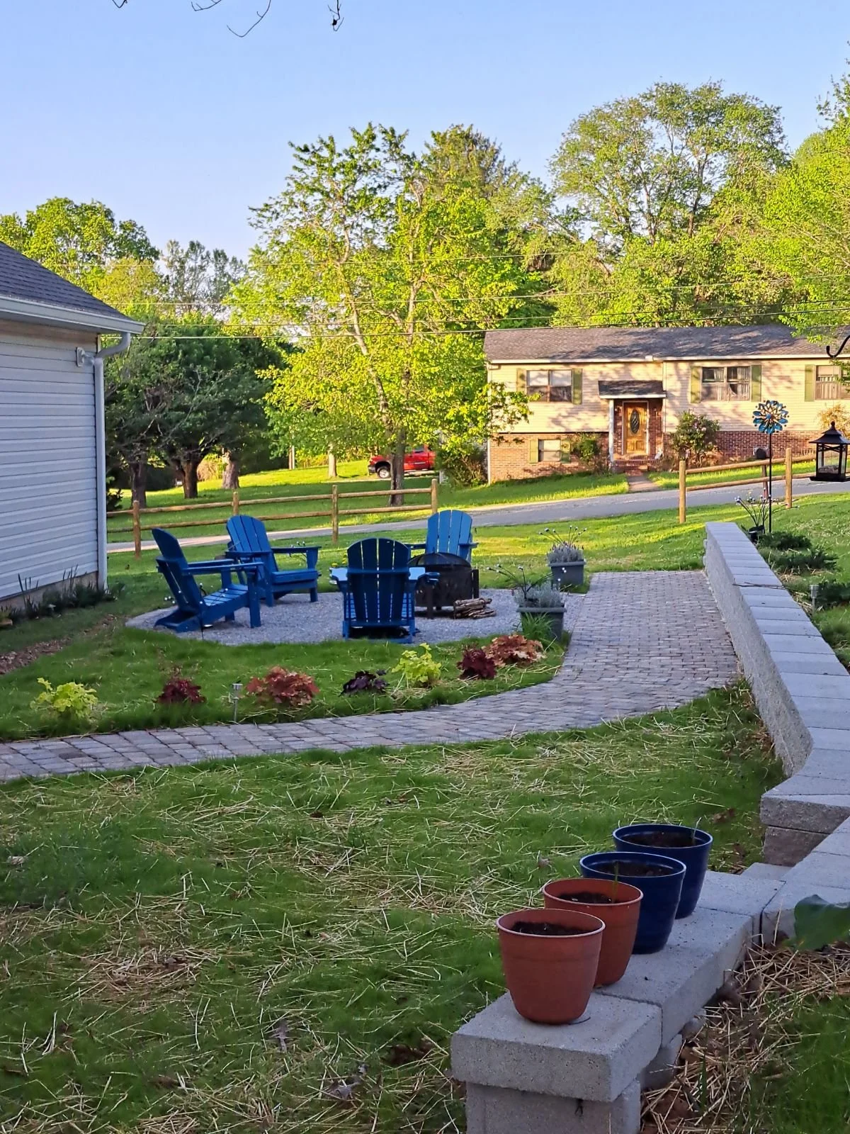 Backyard garden with a brick pathway, colorful potted plants, a fire pit surrounded by blue Adirondack chairs, a white fence, green grass, trees, and a house in the background.