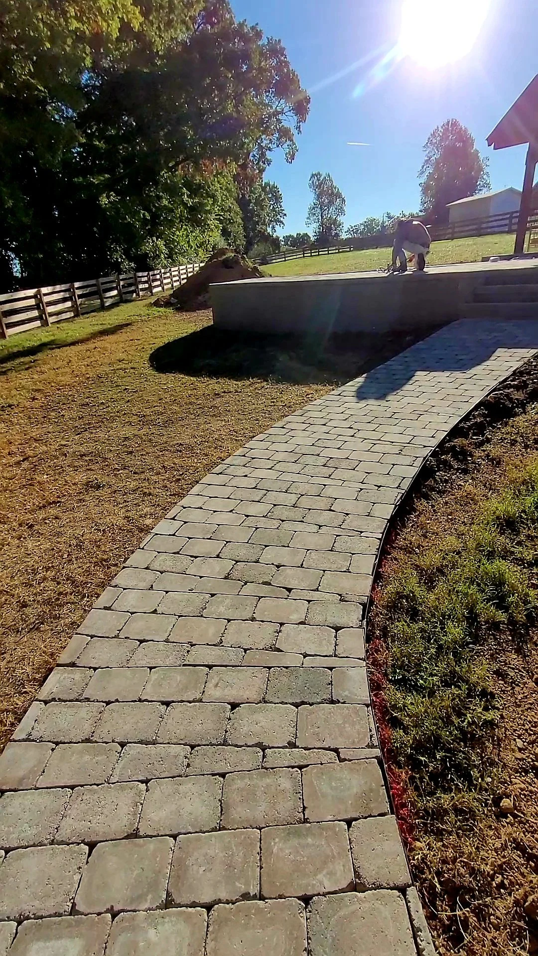 A worker installing brick pavers on a curved pathway in a landscaped yard with a fence and trees in the background, bathed in sunlight.