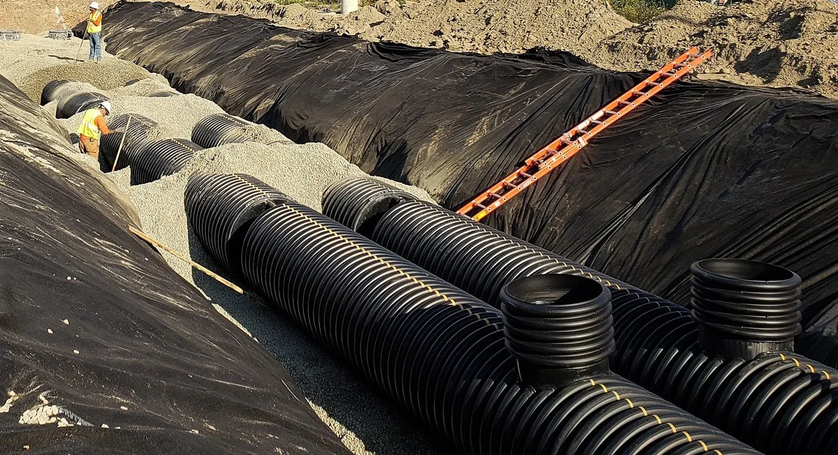 Construction workers installing large black corrugated plastic pipes in an excavated trench for drainage or sewer system.