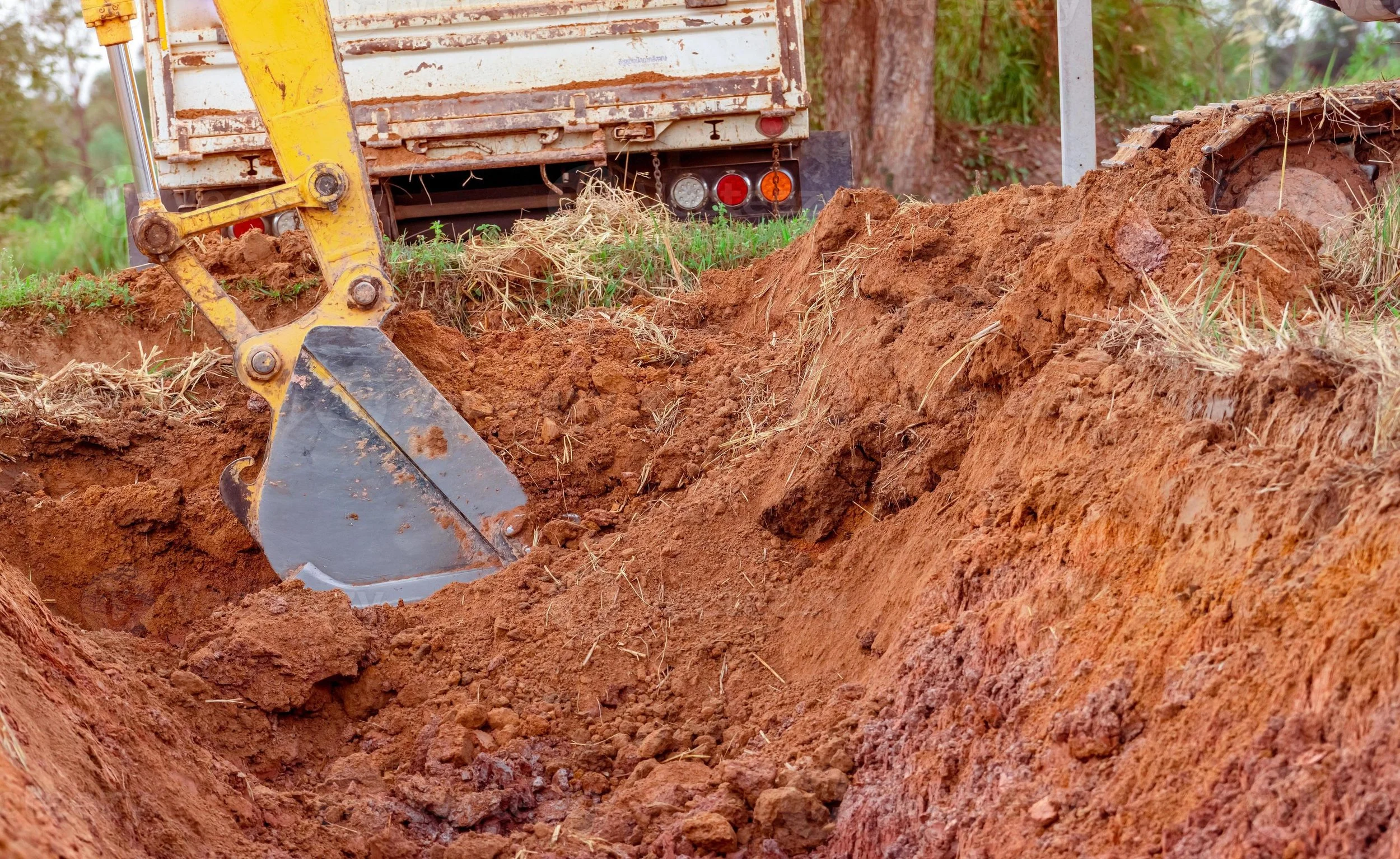 A construction site with a yellow excavator digging a trench in the ground, with dirt and grass around.