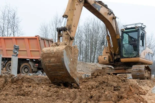 Excavator working on a construction site, digging into dirt with a dump truck nearby.