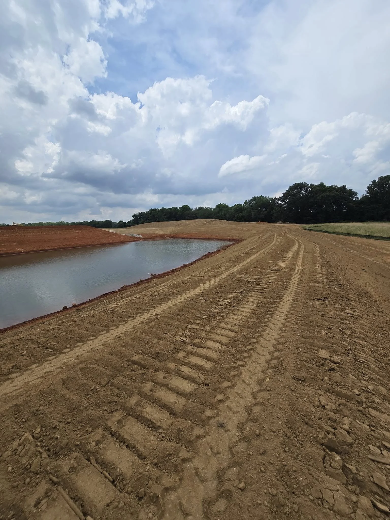 A dirt road with tire tracks runs alongside a small pond with a dirt embankment, with green trees in the background under a sky filled with clouds.