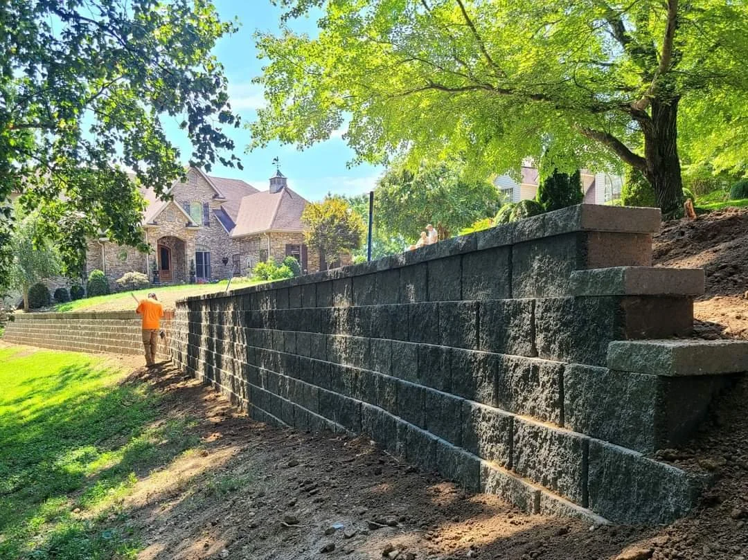 A worker standing next to a newly built stone retaining wall in a residential backyard, with a large tree providing shade and a house under construction in the background.