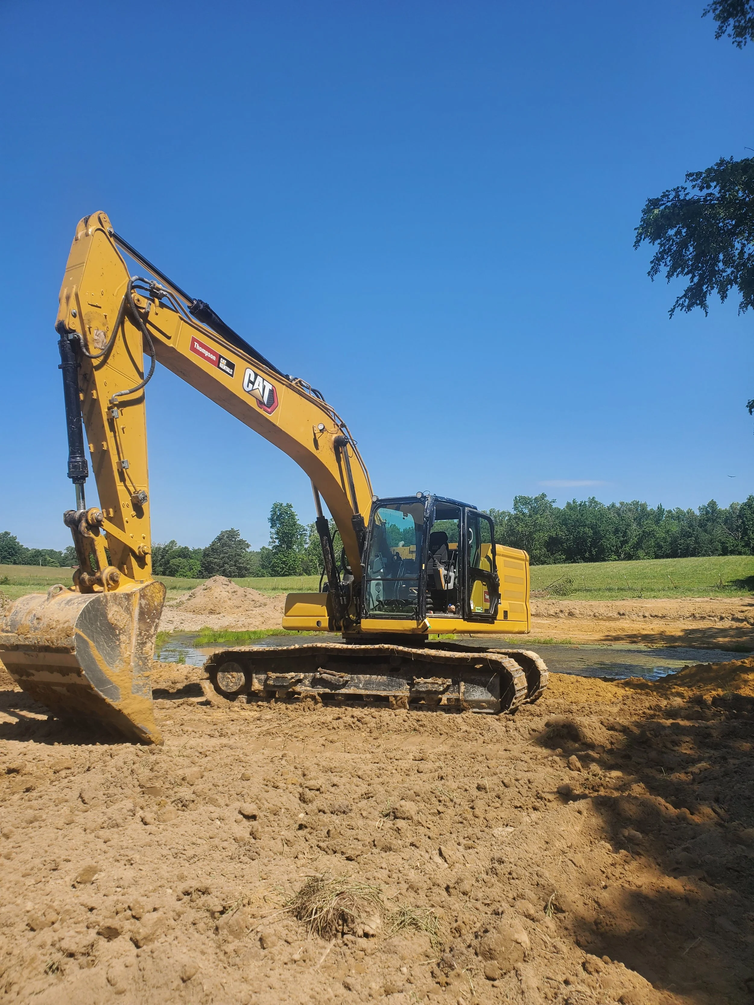 A yellow excavator with tracked wheels is working on a construction site under a clear blue sky and some trees in the background.