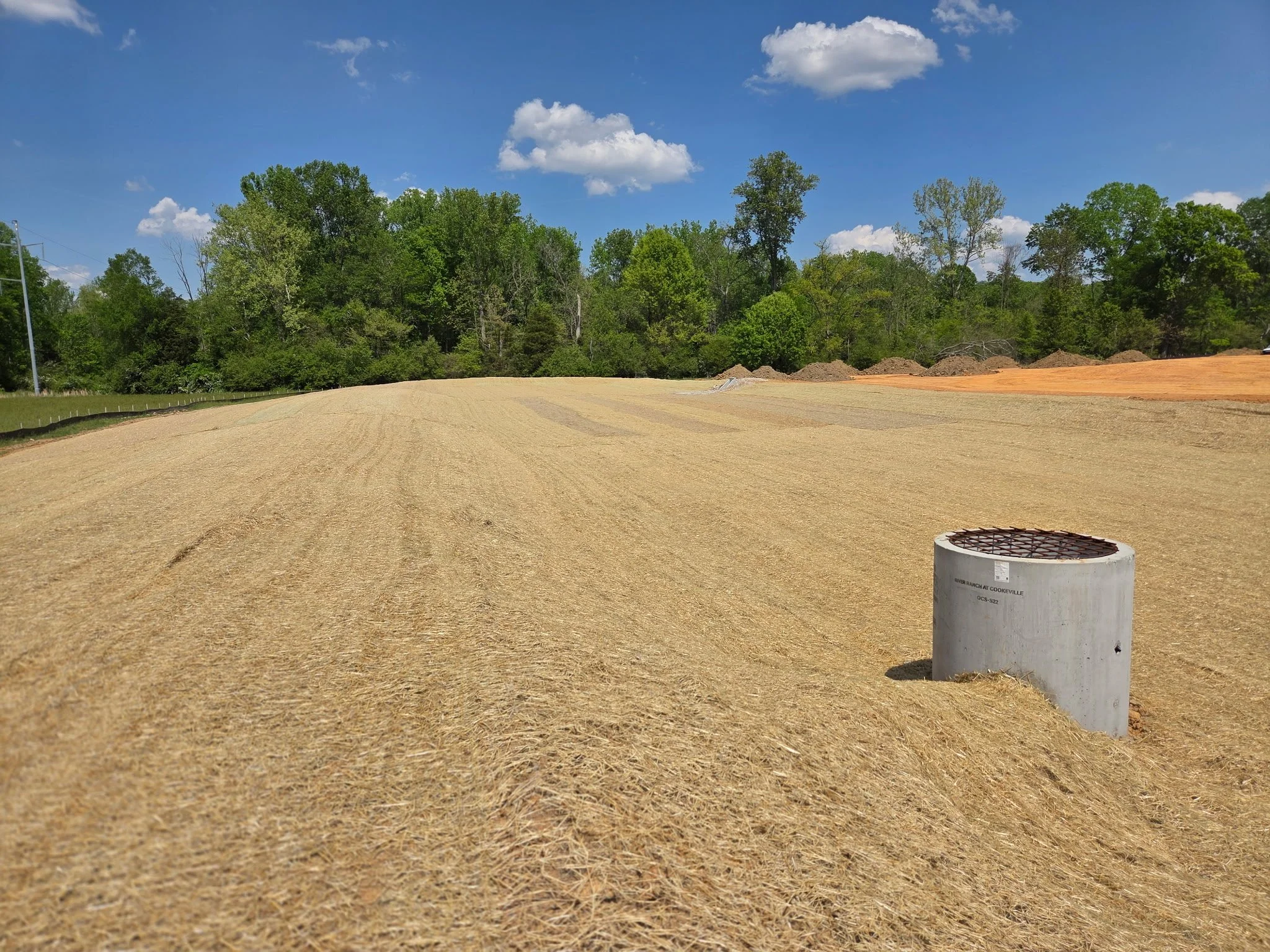 A field with light brown soil and a row of trees with green leaves in the background under a blue sky with fluffy white clouds. There is a metal pipe cap with a label and a small ladder or ramp in the bottom right corner.