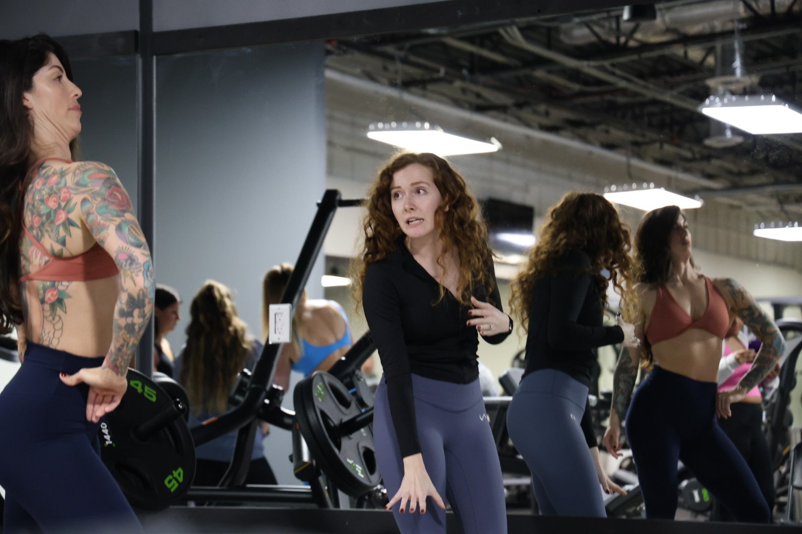 Group of women participating in a fitness class at a gym, standing in front of mirrors, with one woman in the center speaking to the group.