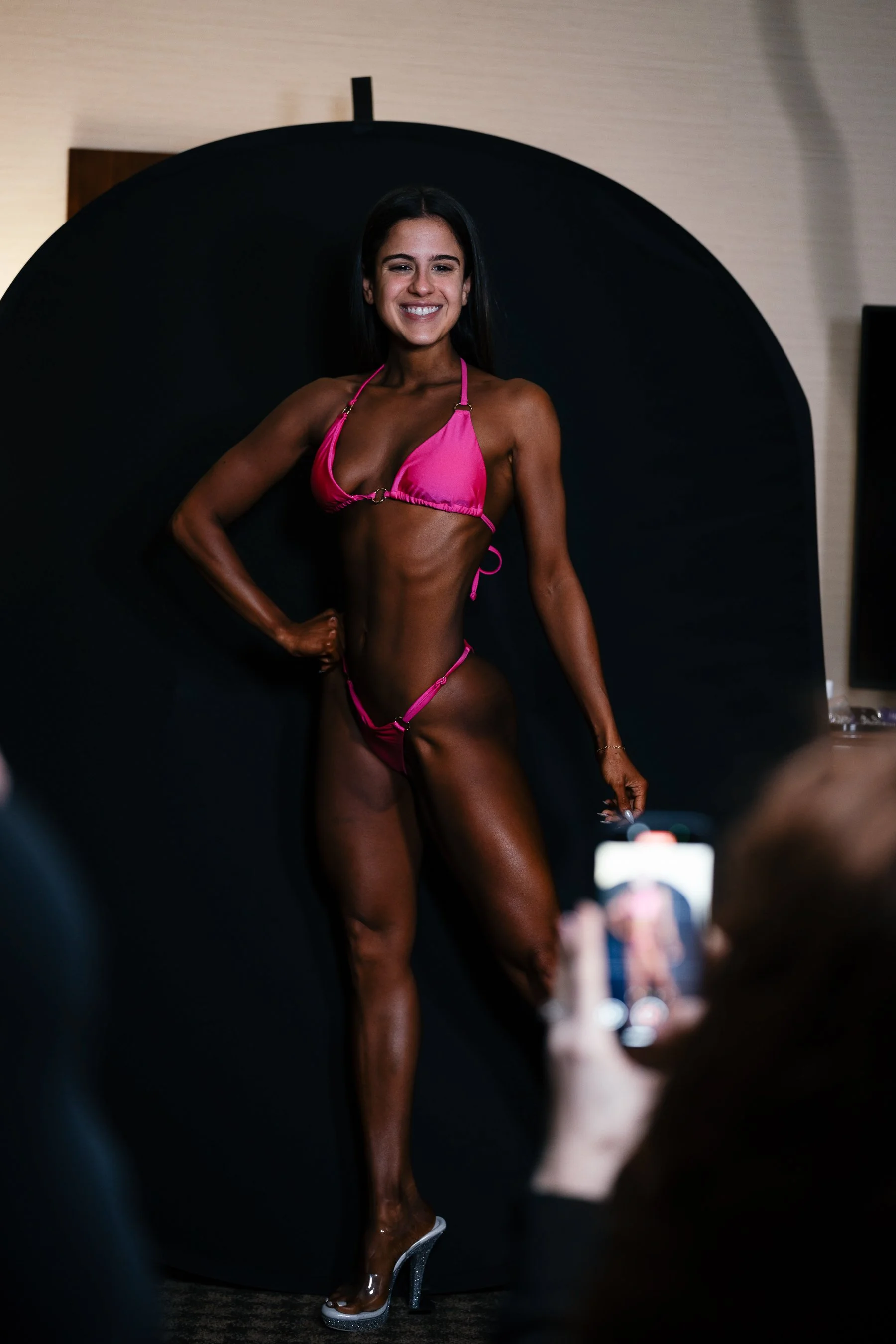 A woman on stage wearing a bikini with long blonde hair, posing with her back to the camera. The background shows a banner with the words 'Atlantic Coast Championship 2025' and 'Bikini'.