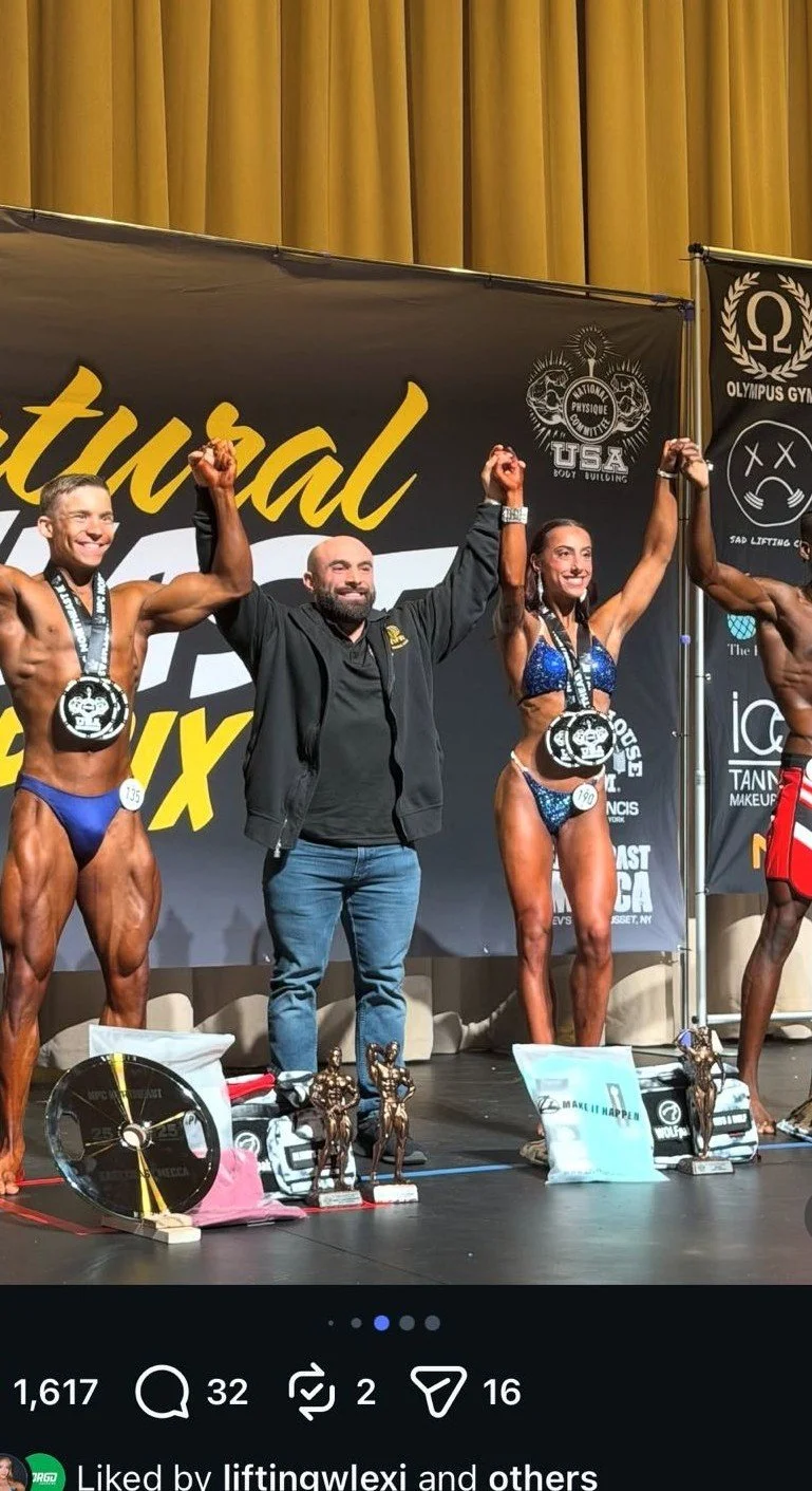 Two female bodybuilders and a male judge or organizer on a stage at a bodybuilding competition, holding hands raised in victory, with medals and trophies in front of them.