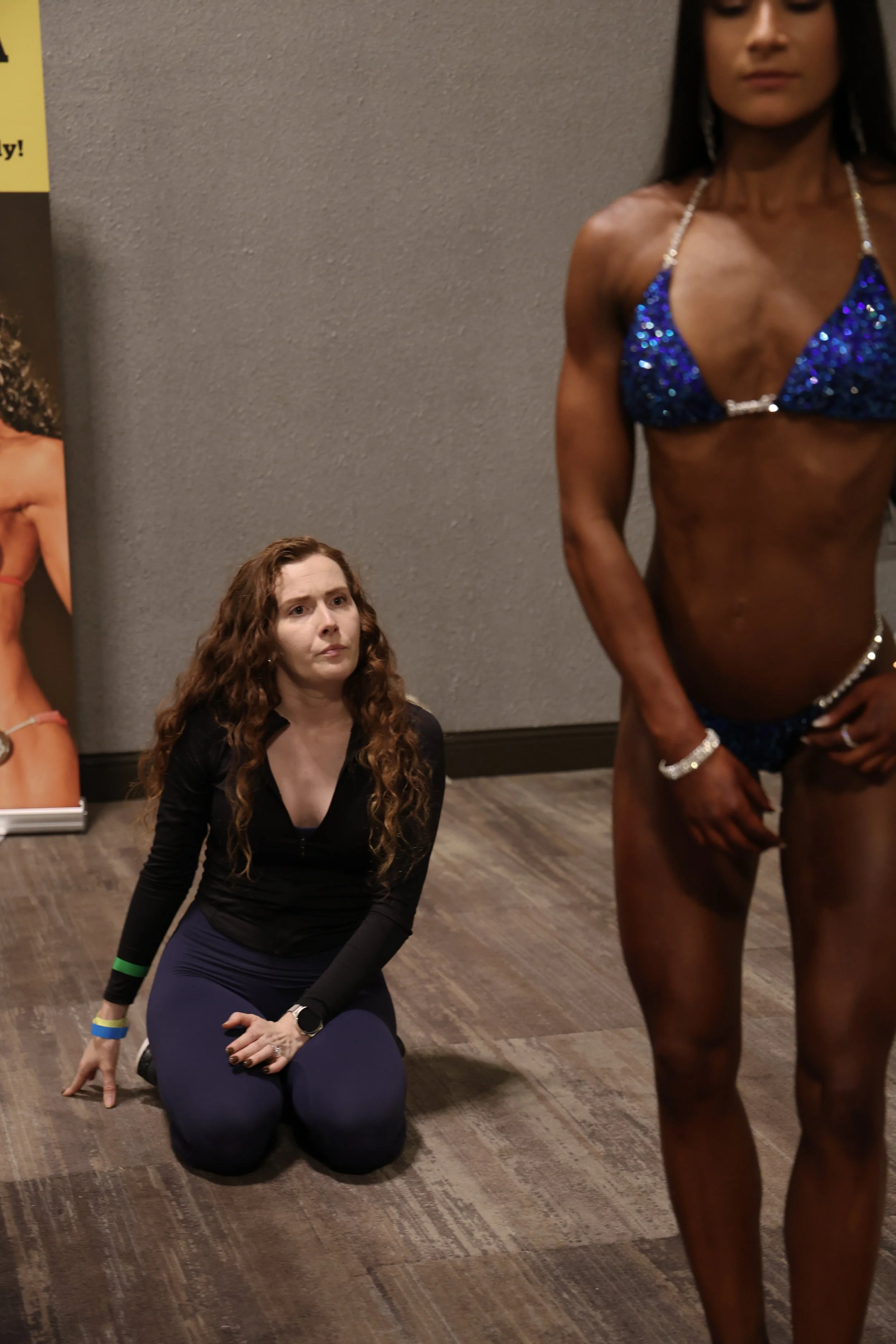 A woman with long red hair is kneeling on the floor with a serious expression, observing a muscular woman in a blue bikini showing her physique at a fitness or bodybuilding event.