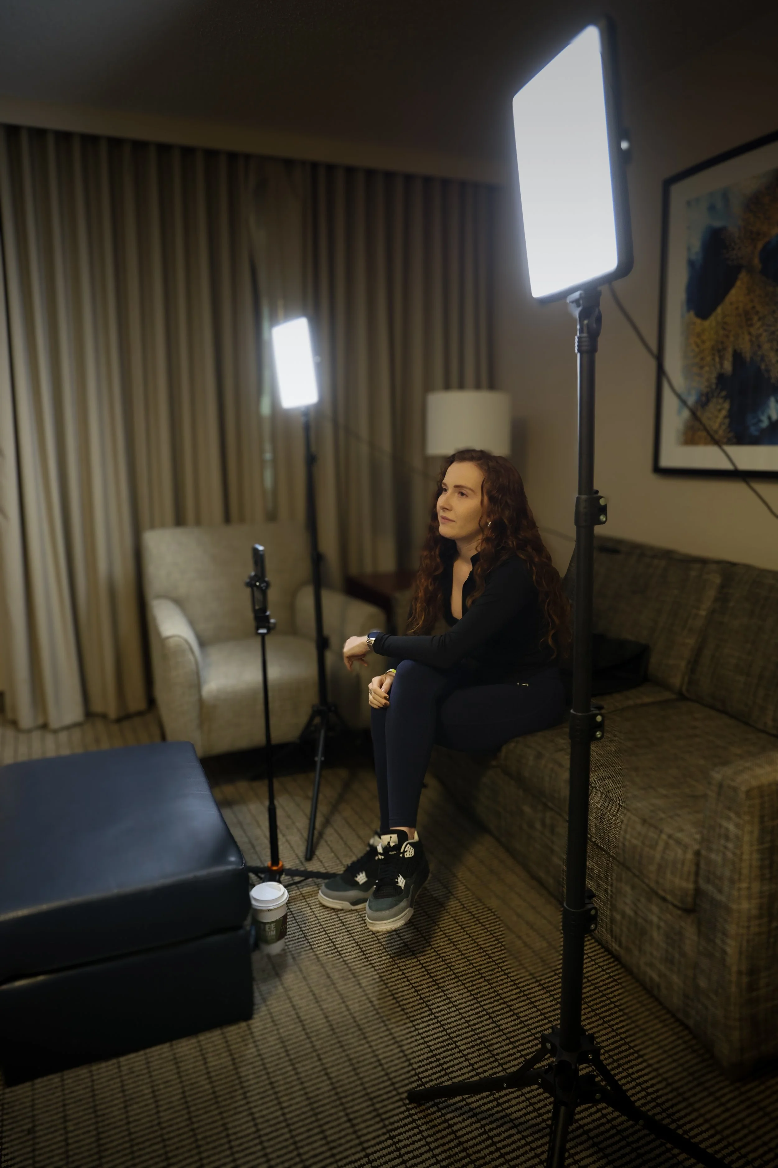 Woman with curly hair sitting on a couch, being filmed with studio lights in a room with curtains and a framed picture on the wall.