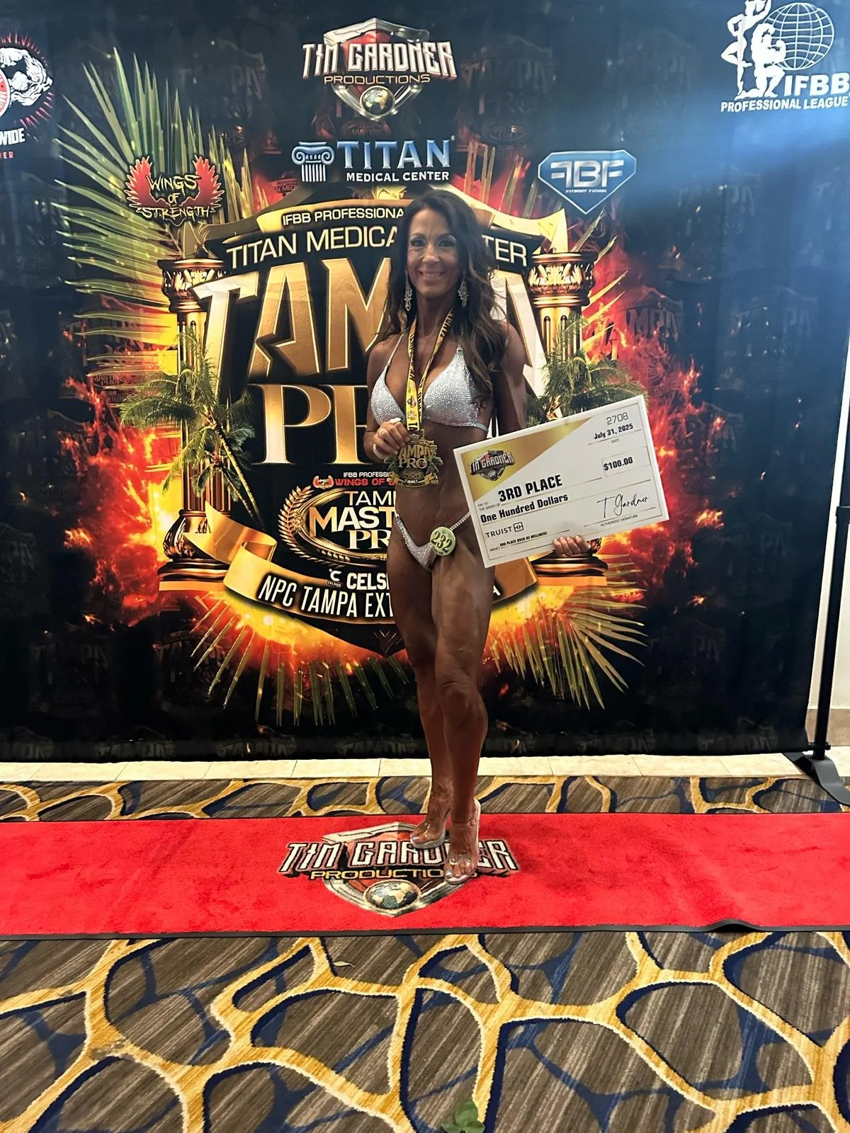 A woman in a silver bikini holding a large check and a medal, standing on a red carpet, smiling at a bodybuilding competition awards ceremony.