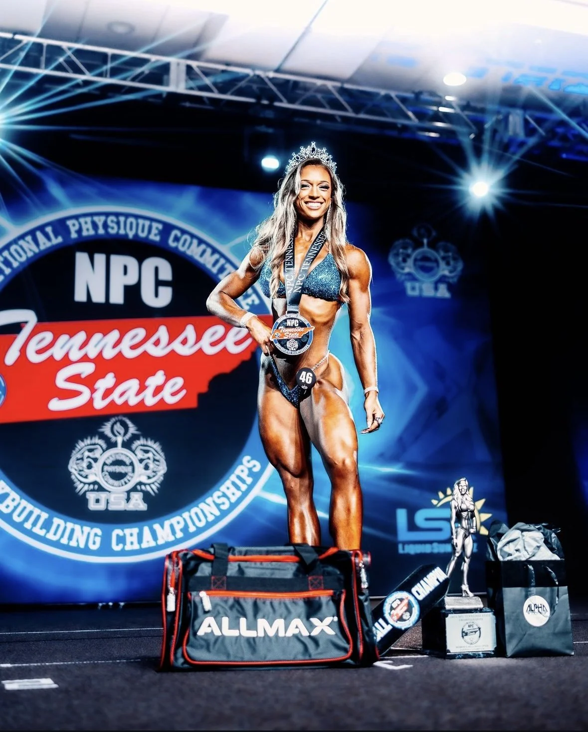 A female bodybuilder on stage at a competition, wearing a sparkly bikini and a crown, holding medals, with trophies and bags in front of her. The backdrop features the logo of the NPC Tennessee State Championships.