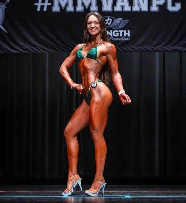 A female bodybuilder on stage during a competition, wearing a sparkly green bikini and high heels, posing with her left arm bent and her right arm on her hip, smiling at the camera.