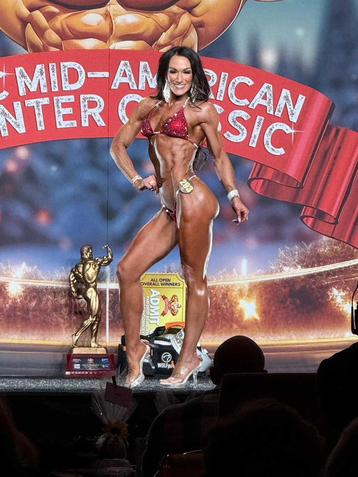 A female bodybuilder on stage at a bodybuilding competition, wearing a red bikini and high heels, striking a pose beside a trophy.